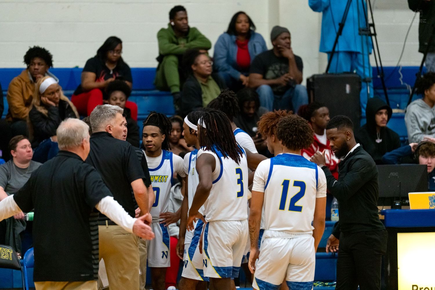 Northwest Knights basketball players and coaches gather during a timeout in a packed gymnasium.