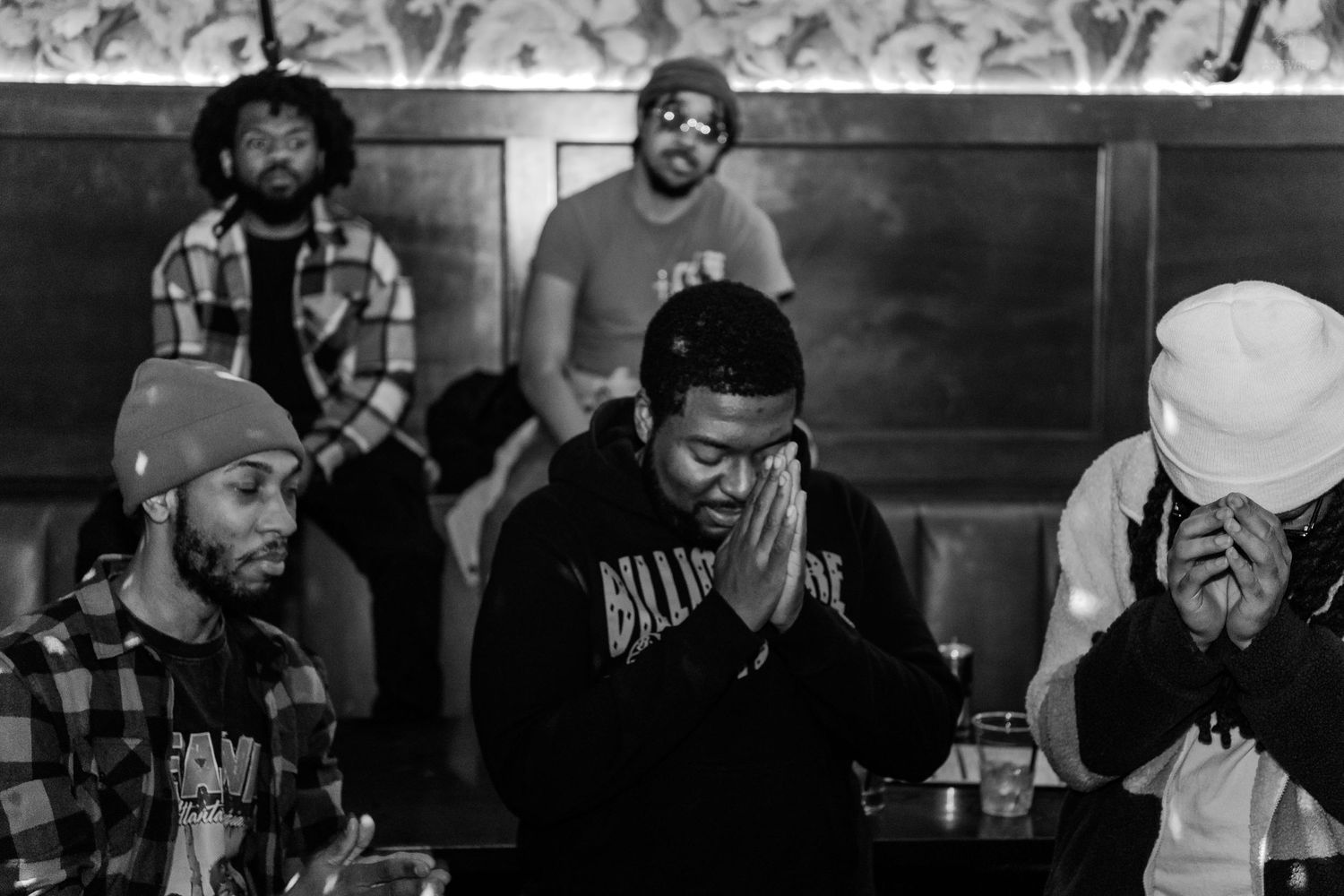 Black and white photo of a group of men with hands clasped in prayer pose at an indoor venue.