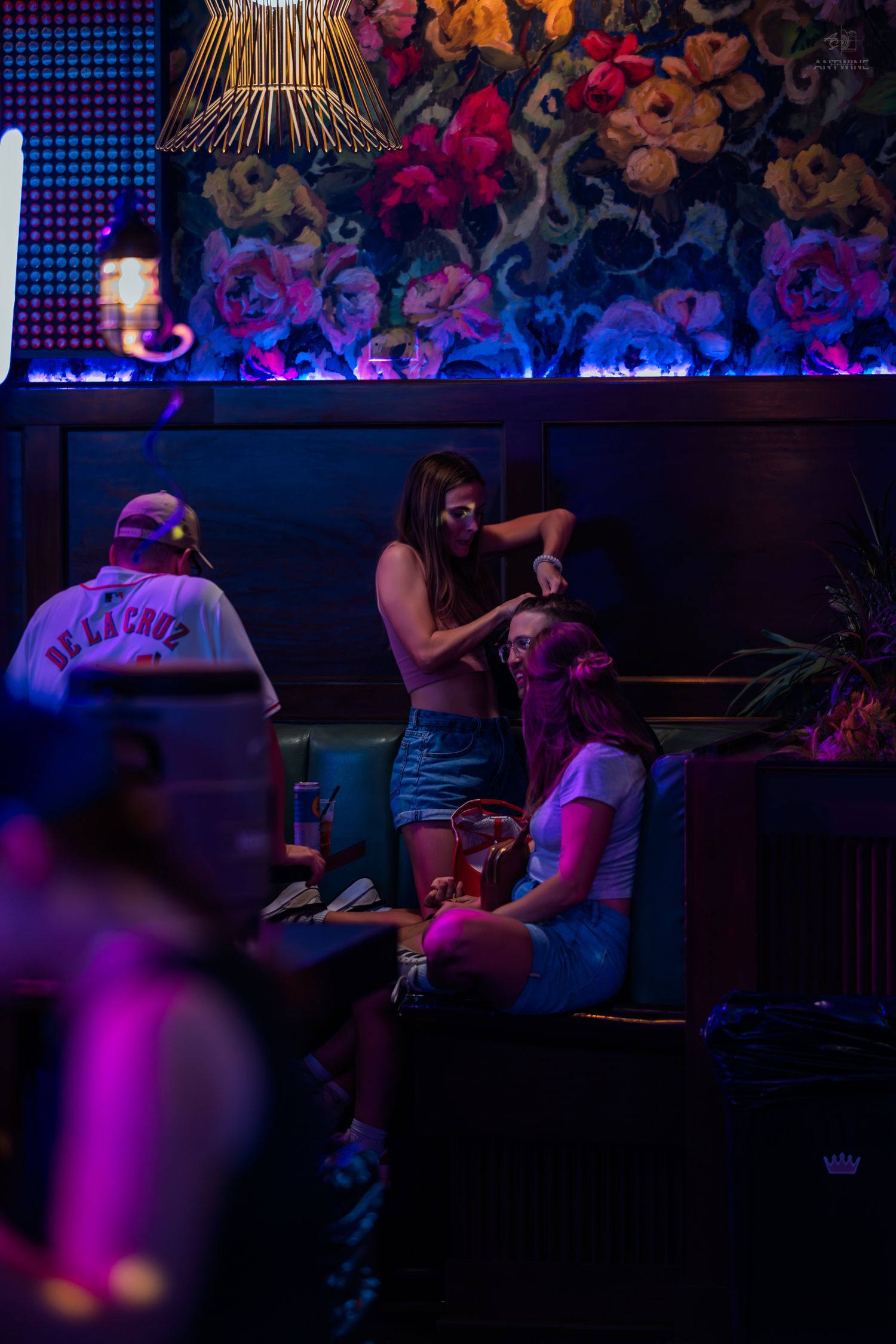 Young women socializing in a neon-lit bar booth with floral wall decor.