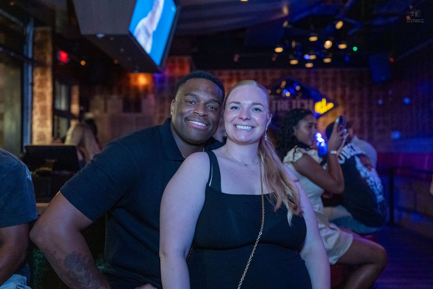 A smiling couple poses together at a lively bar, with colorful lights in the background.