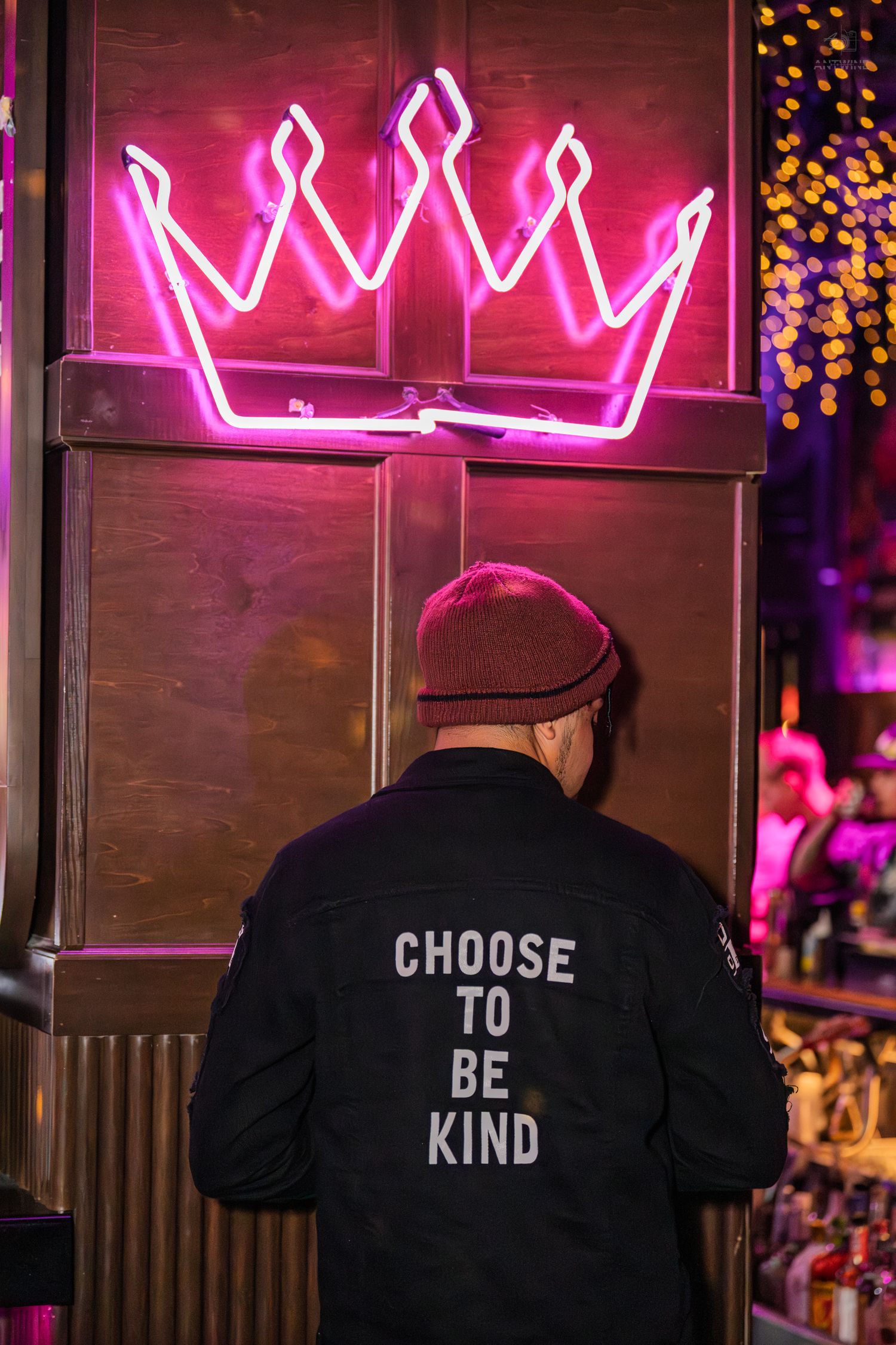 Man in Choose To Be Kind jacket facing a glowing pink neon crown at a bar.