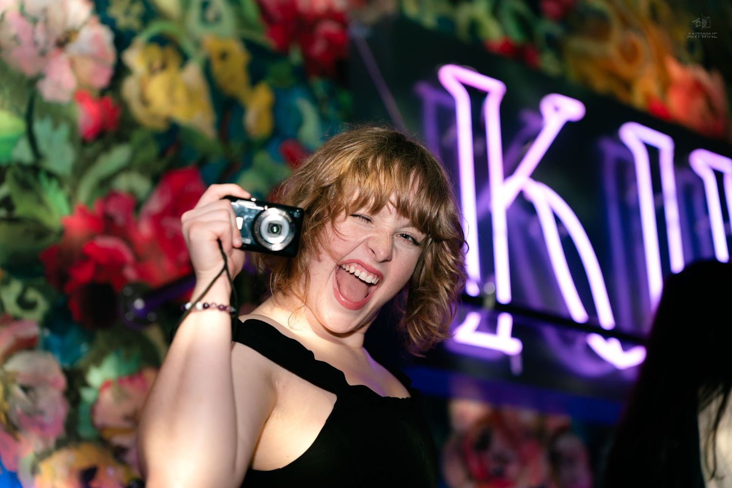 Laughing woman holding a compact camera near a glowing floral neon K sign.