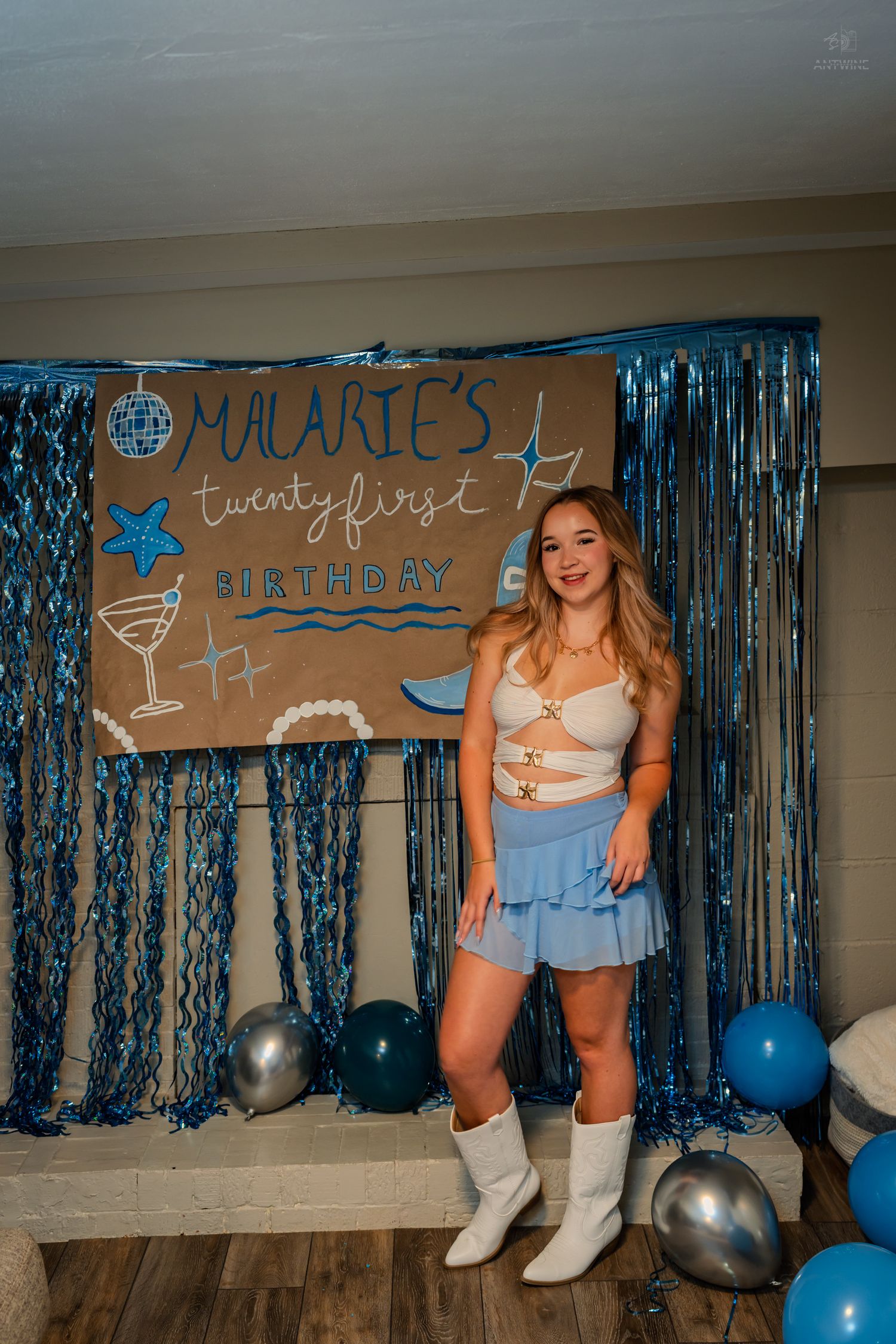 Smiling young woman posing in front of handmade Malarie's 21st birthday sign with blue balloons.