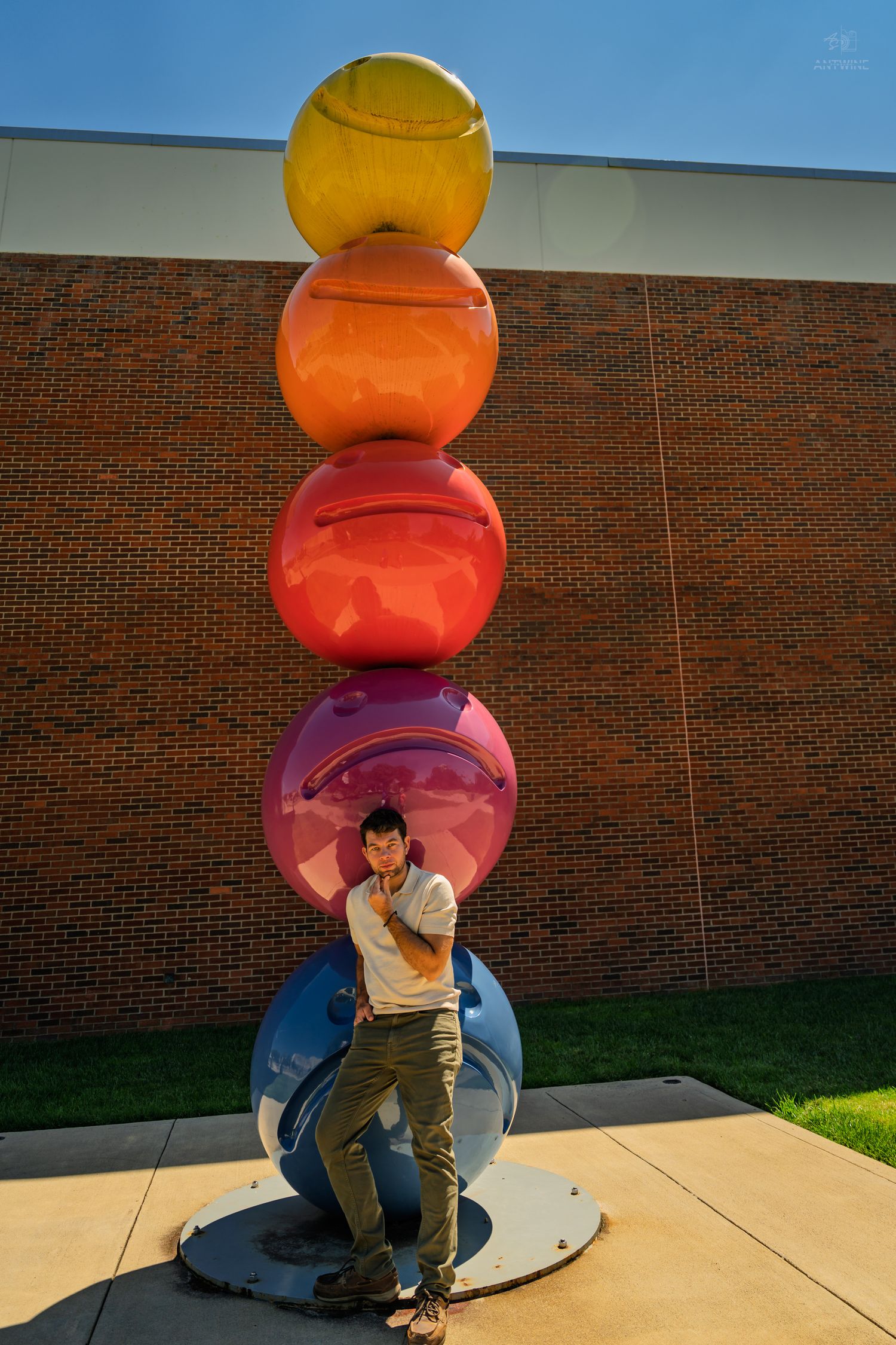 Man in casual outfit leaning against vibrant rainbow stacked spheres sculpture by brick building.