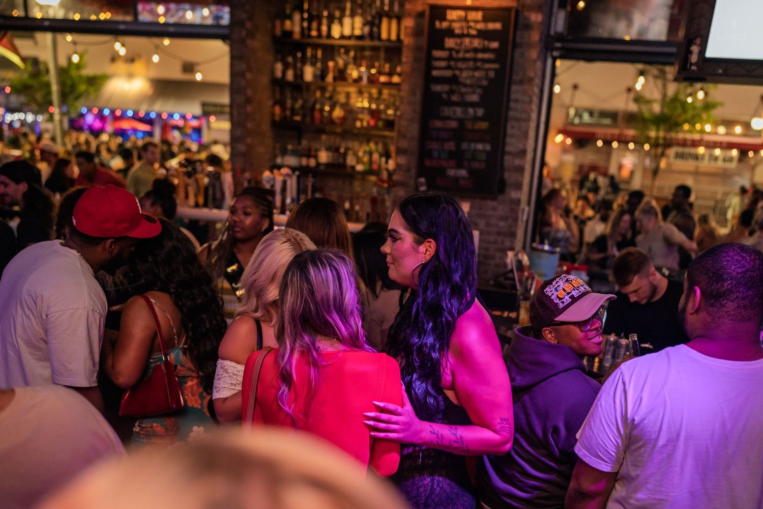 Lively bar crowd with women in colorful outfits socializing under purple lights with bottles displayed.