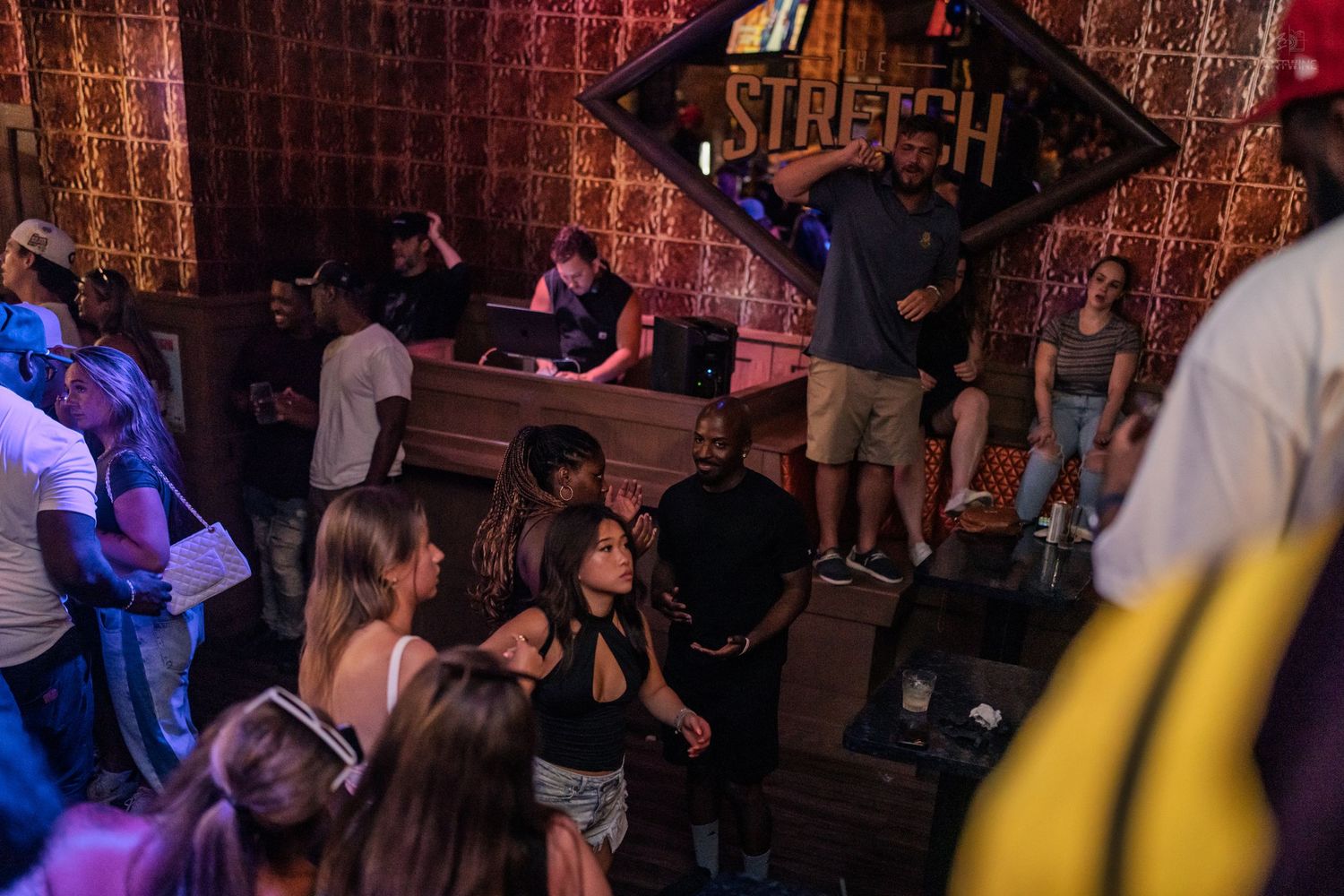 Crowded nightclub with DJ booth, dancers, and neon Stretch sign in dimly lit bar.