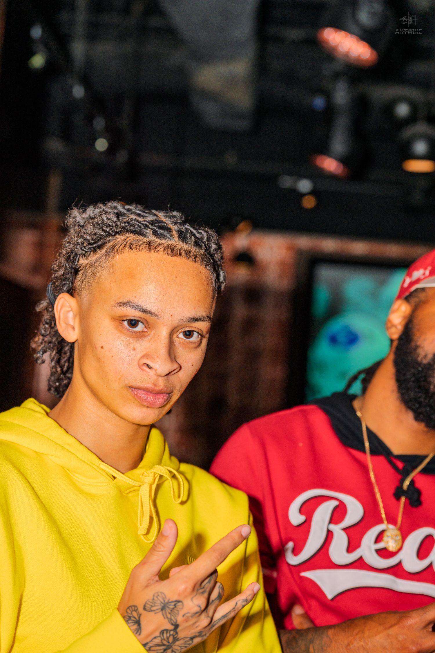 Young person in yellow hoodie making hand gesture at indoor music or nightclub event.