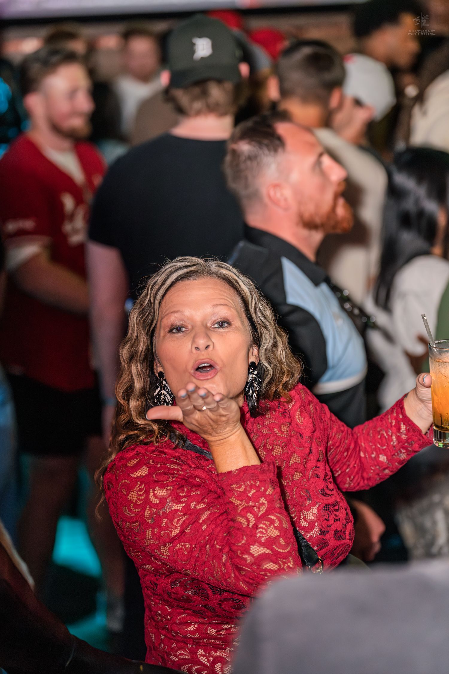 Smiling woman in red lace dress blowing a kiss while holding a cocktail at a bar.