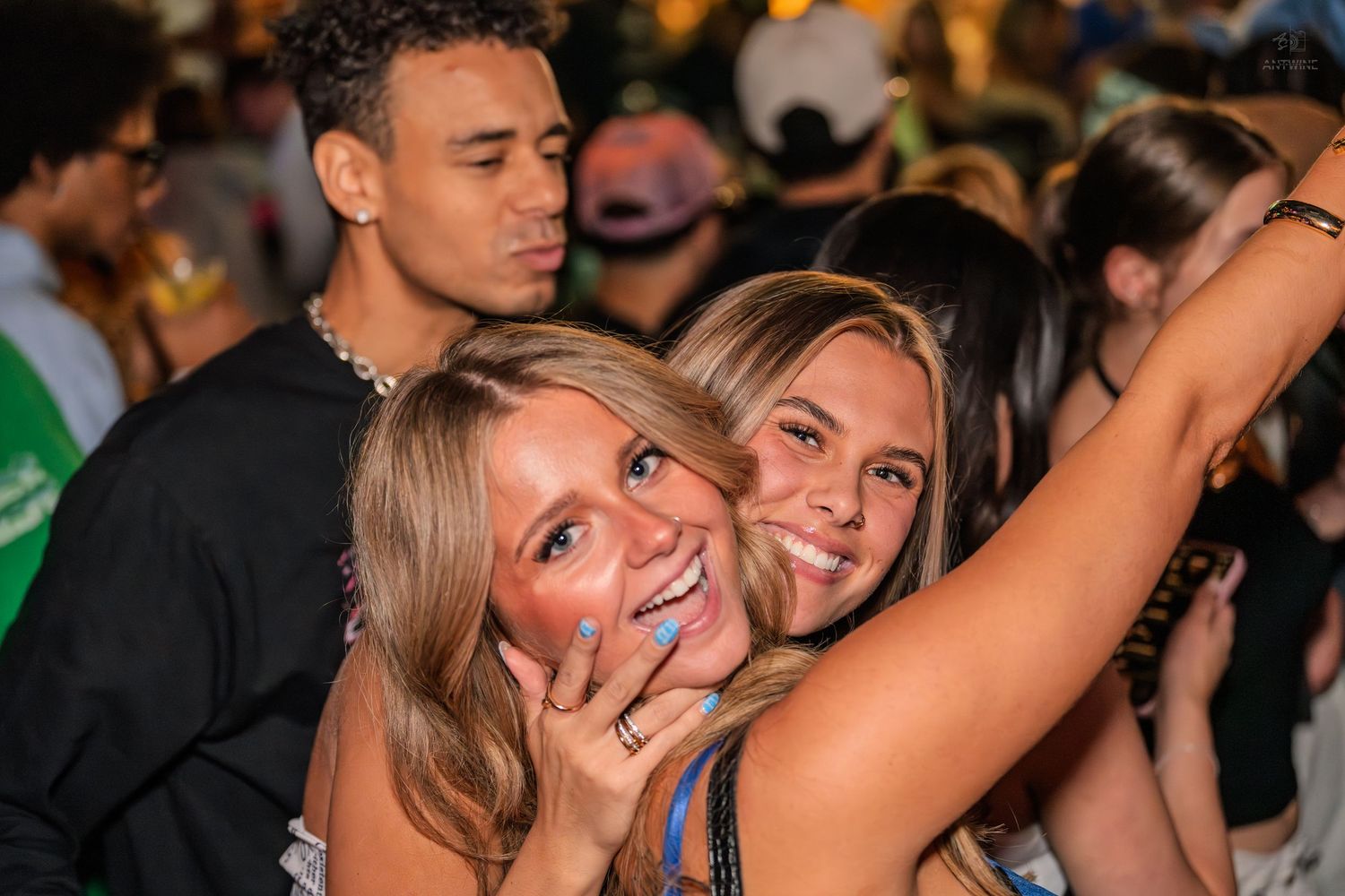 Three young friends smiling and hugging together at a crowded outdoor nighttime social event.