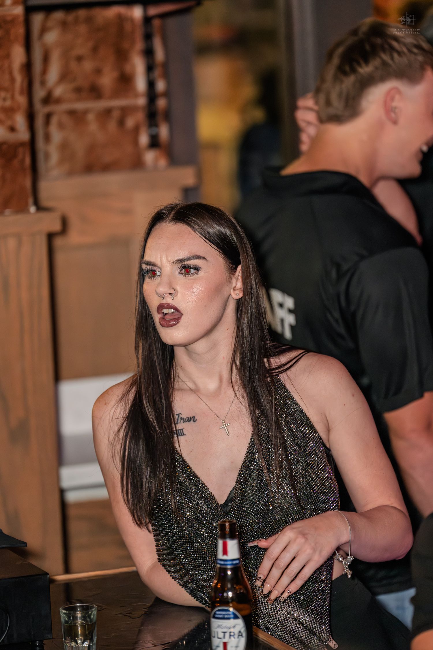 Dark-haired woman in glittery dress holding Michelob Ultra at bar.