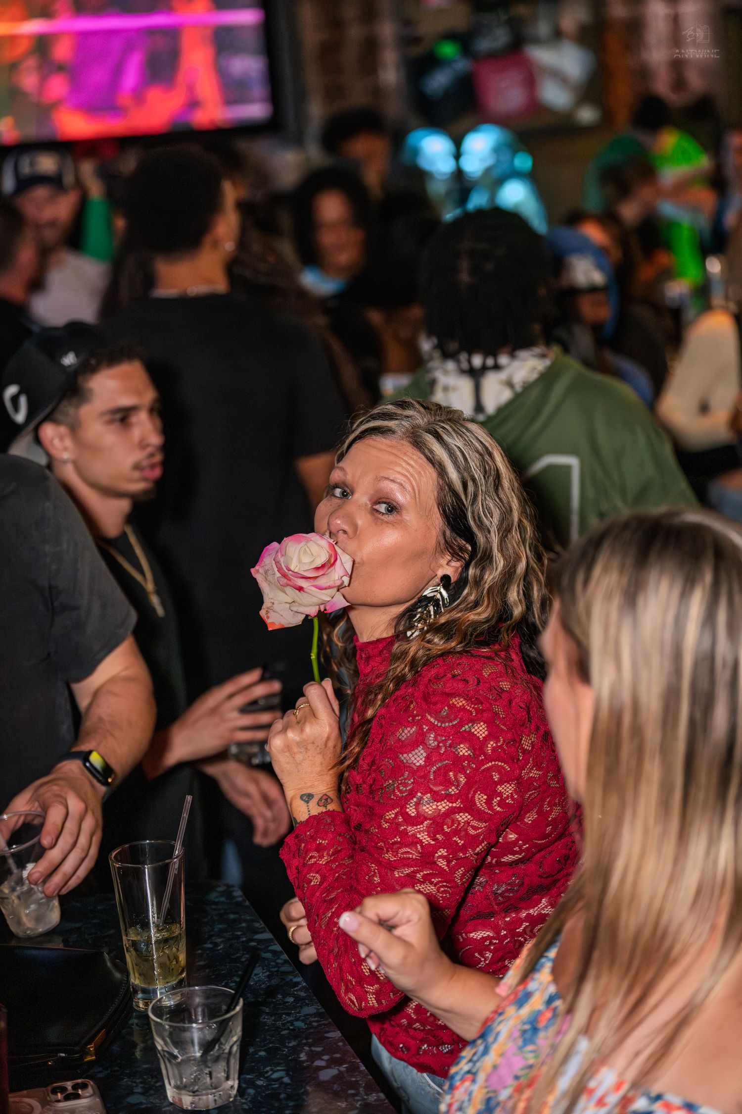 Woman in red lace top playfully holds a pink rose to her lips at a busy nightclub bar.