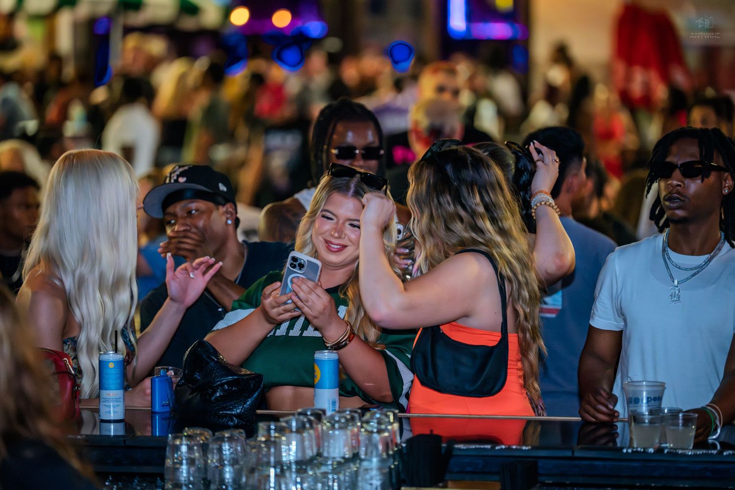 Group of women laughing and taking selfies at a crowded outdoor nightclub bar event.