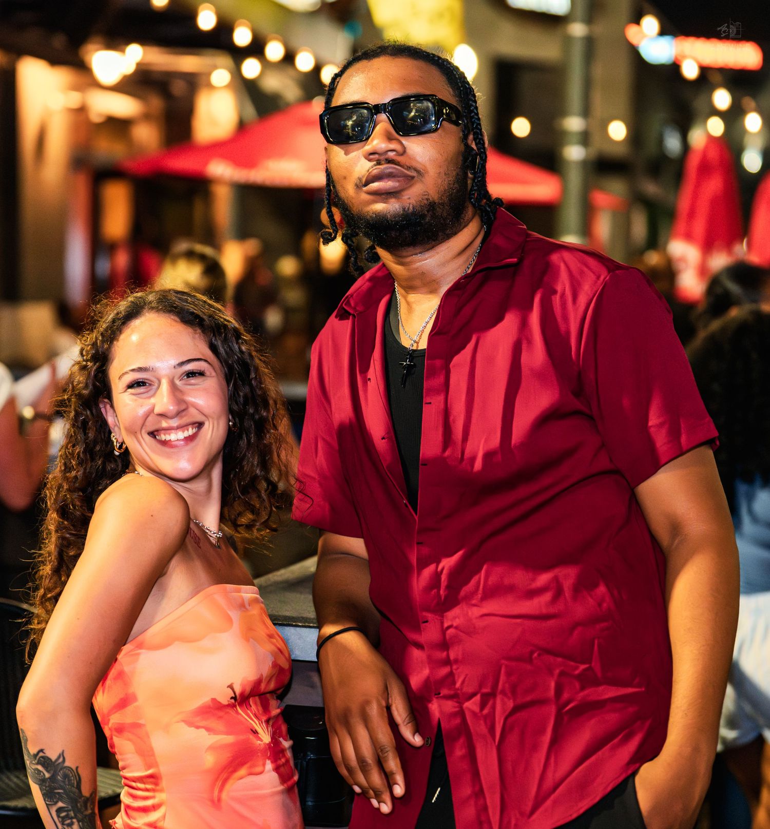 A smiling woman in an orange floral top poses with a man in a red shirt and sunglasses at a nighttime outdoor event.