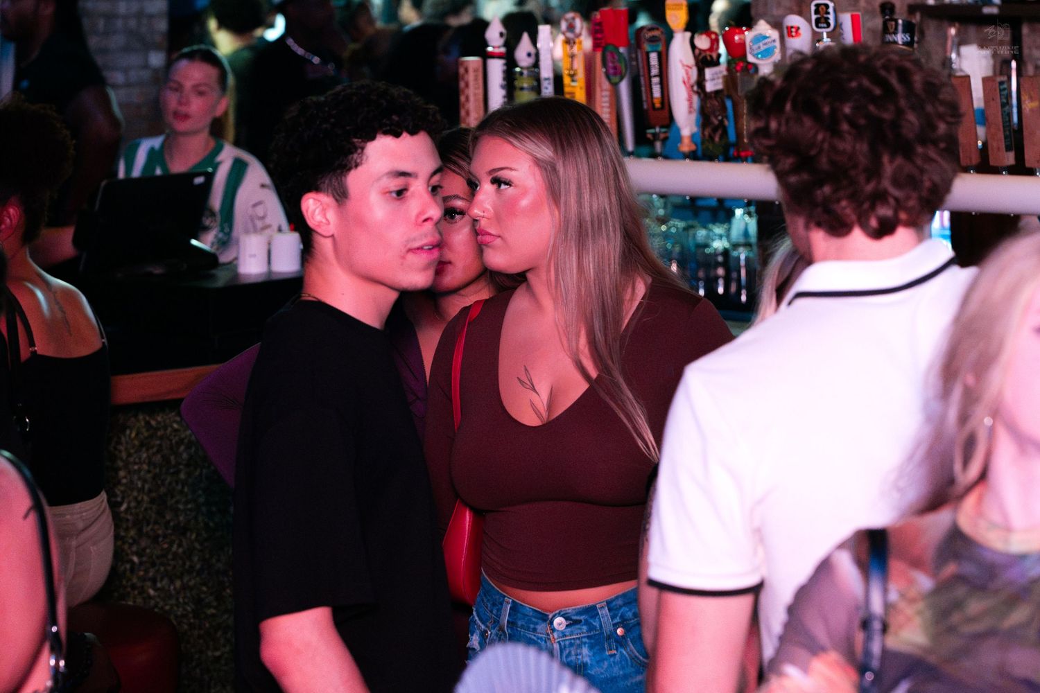 Young couple standing close together near a bar in a busy nightclub setting.