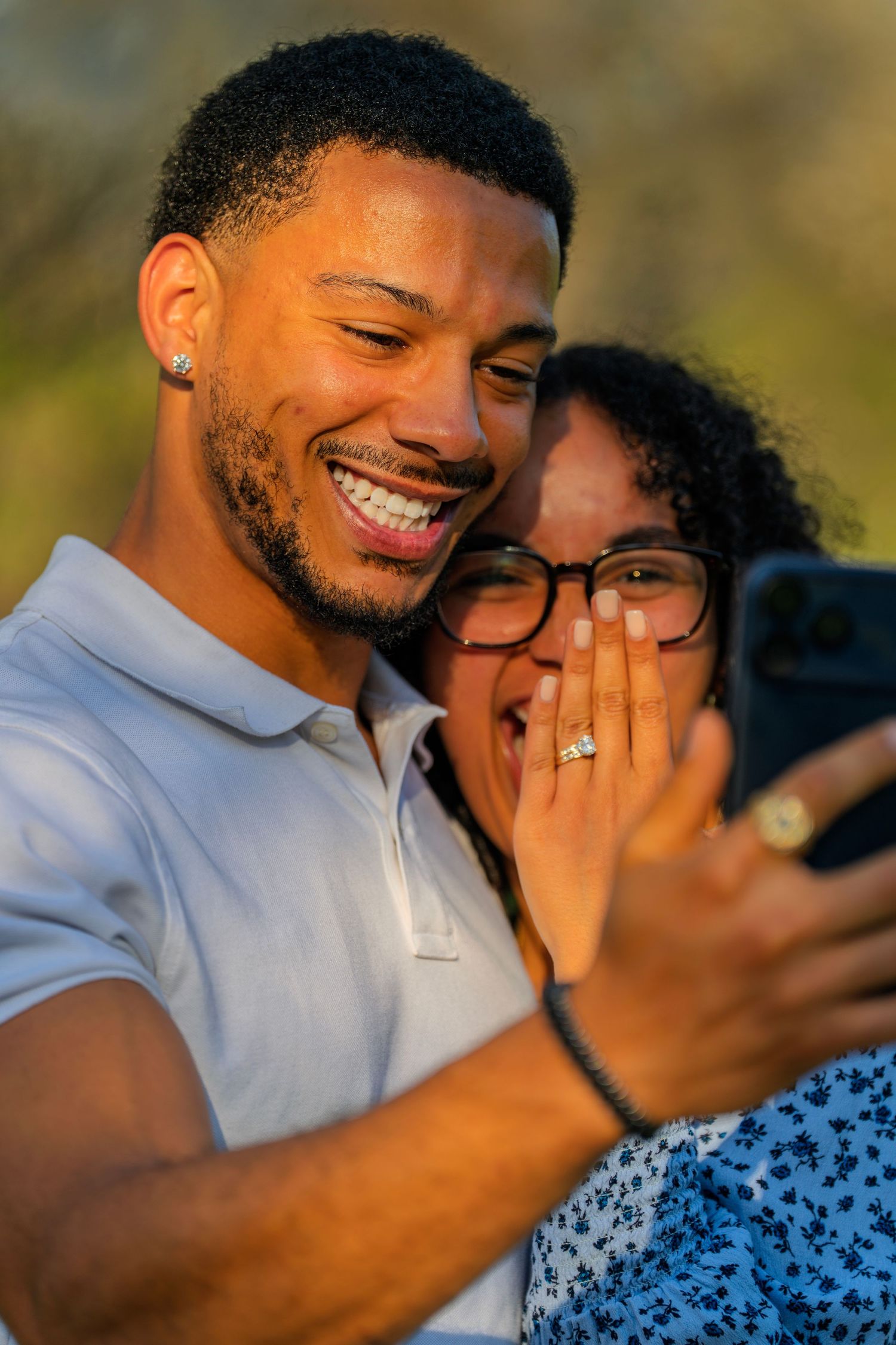 A joyful couple smiles while looking at a smartphone, the woman covering her mouth in surprise outdoors.