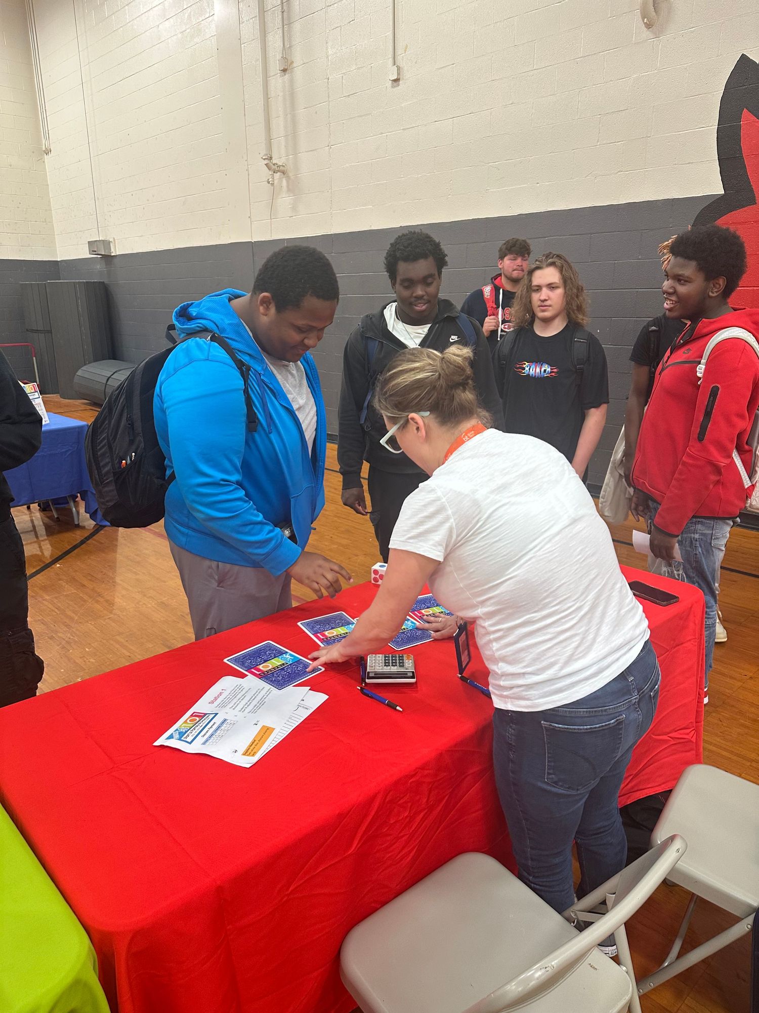 Students speak with a representative at a red table during a school career fair in a gymnasium.