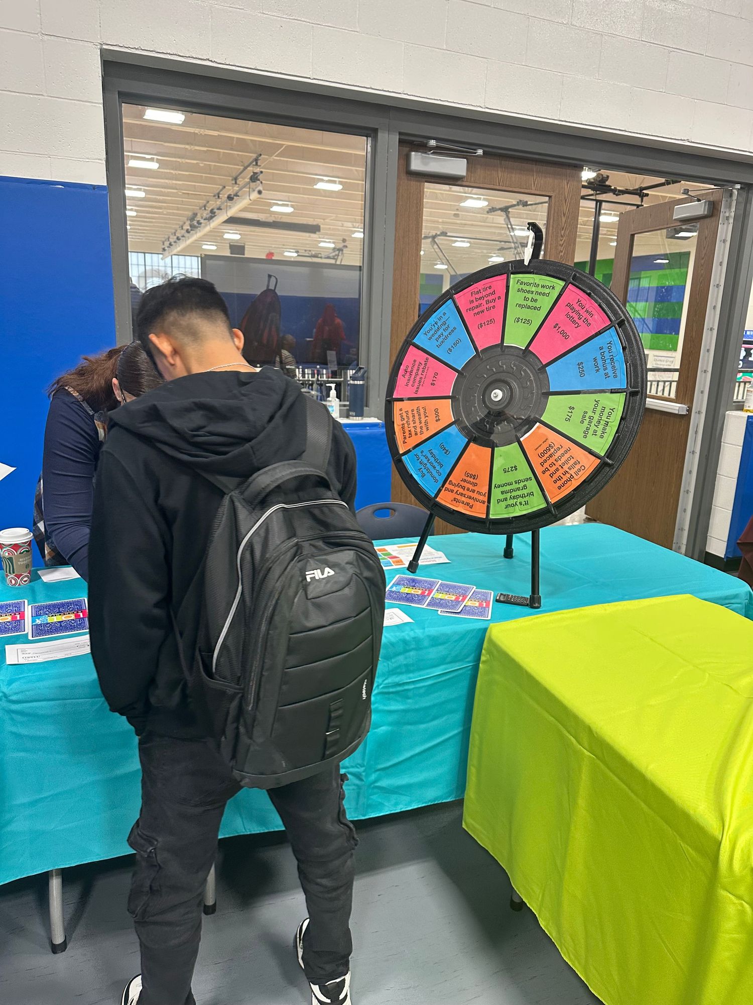 Student with FILA backpack examines a colorful prize wheel at a school fair booth with teal-draped tables.