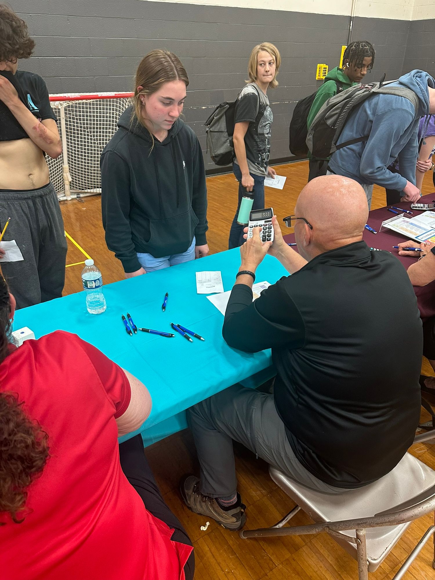 Students consult with a bald male advisor at a blue table during a school guidance fair in a gym.