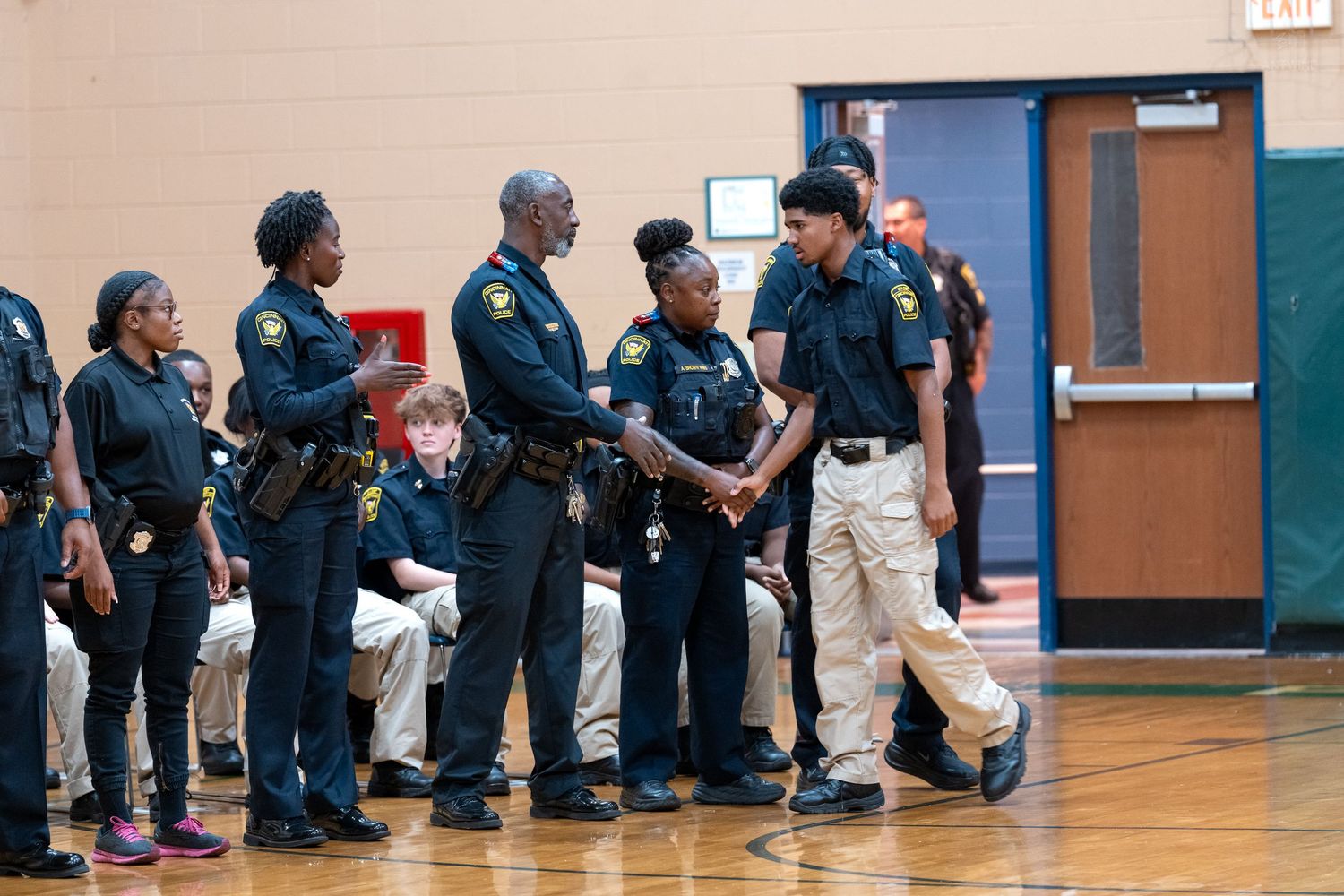 Police officers in uniform shaking hands during a formal recognition ceremony inside a school gymnasium.