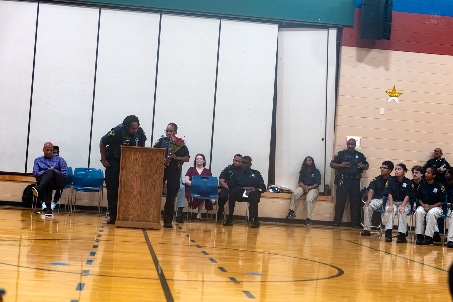 School assembly in gymnasium with speaker at podium, police officers, and students seated along the walls.