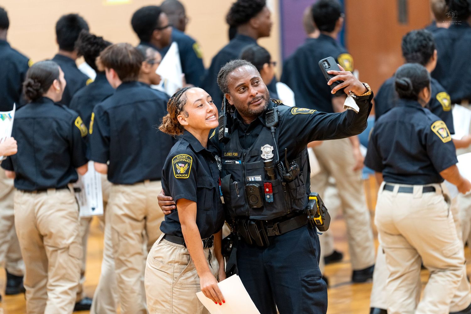 Two police officers in uniform taking a selfie together at an indoor law enforcement graduation ceremony.