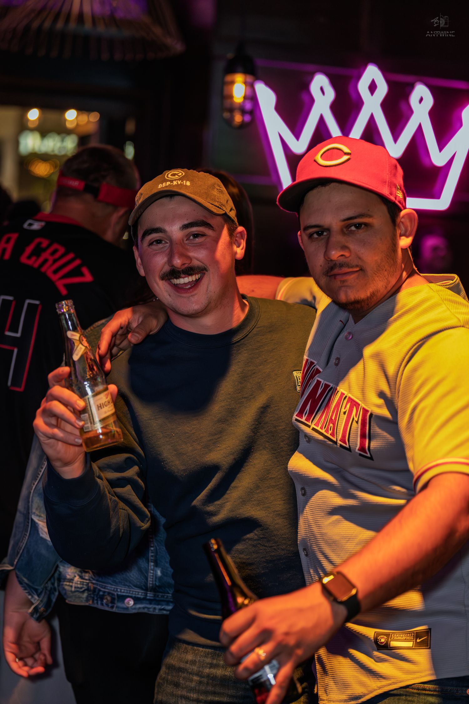 Two friends posing with beers under a glowing pink neon crown at a nightclub.