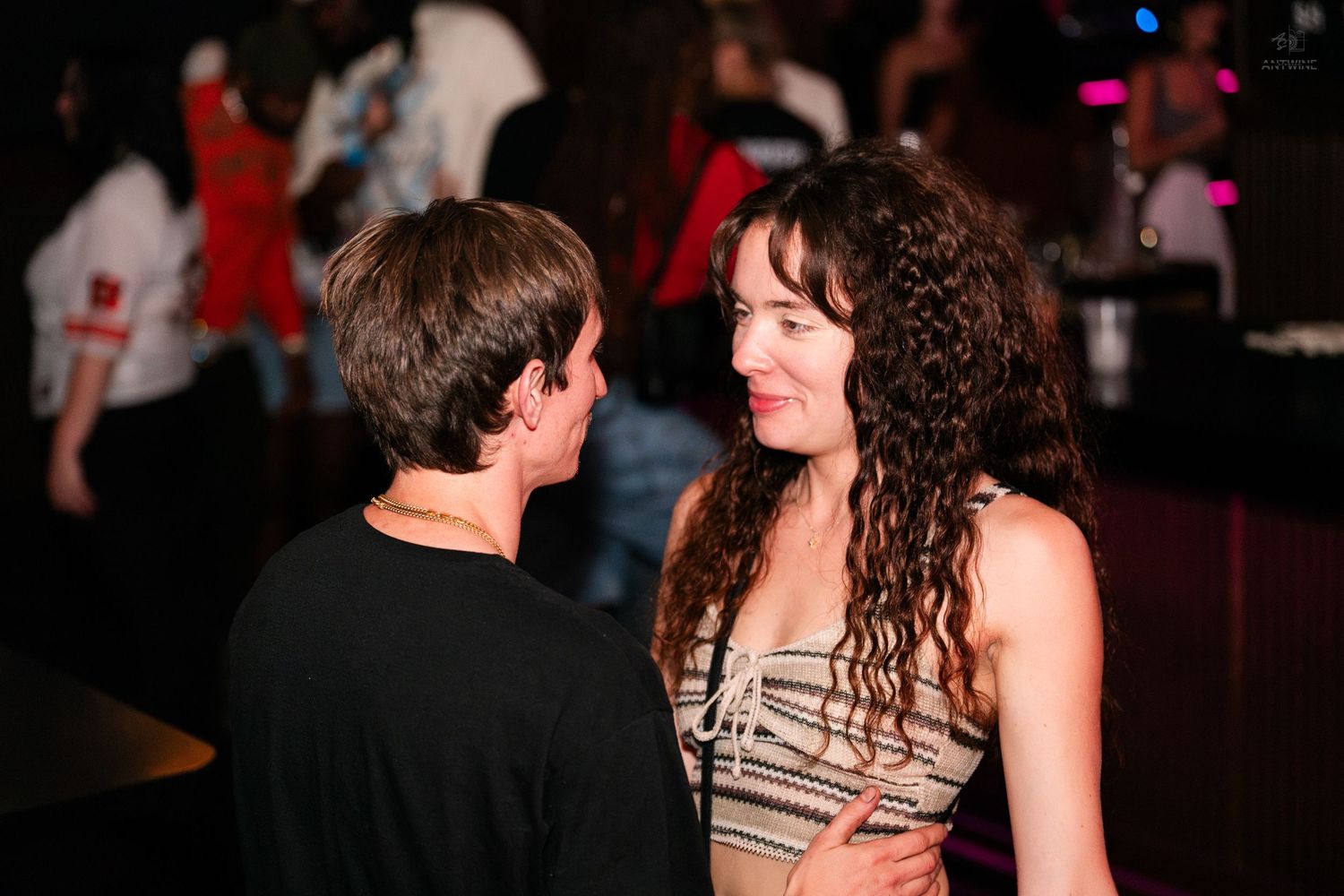 A man and curly-haired woman in a striped top share a moment together on a nightclub dance floor.