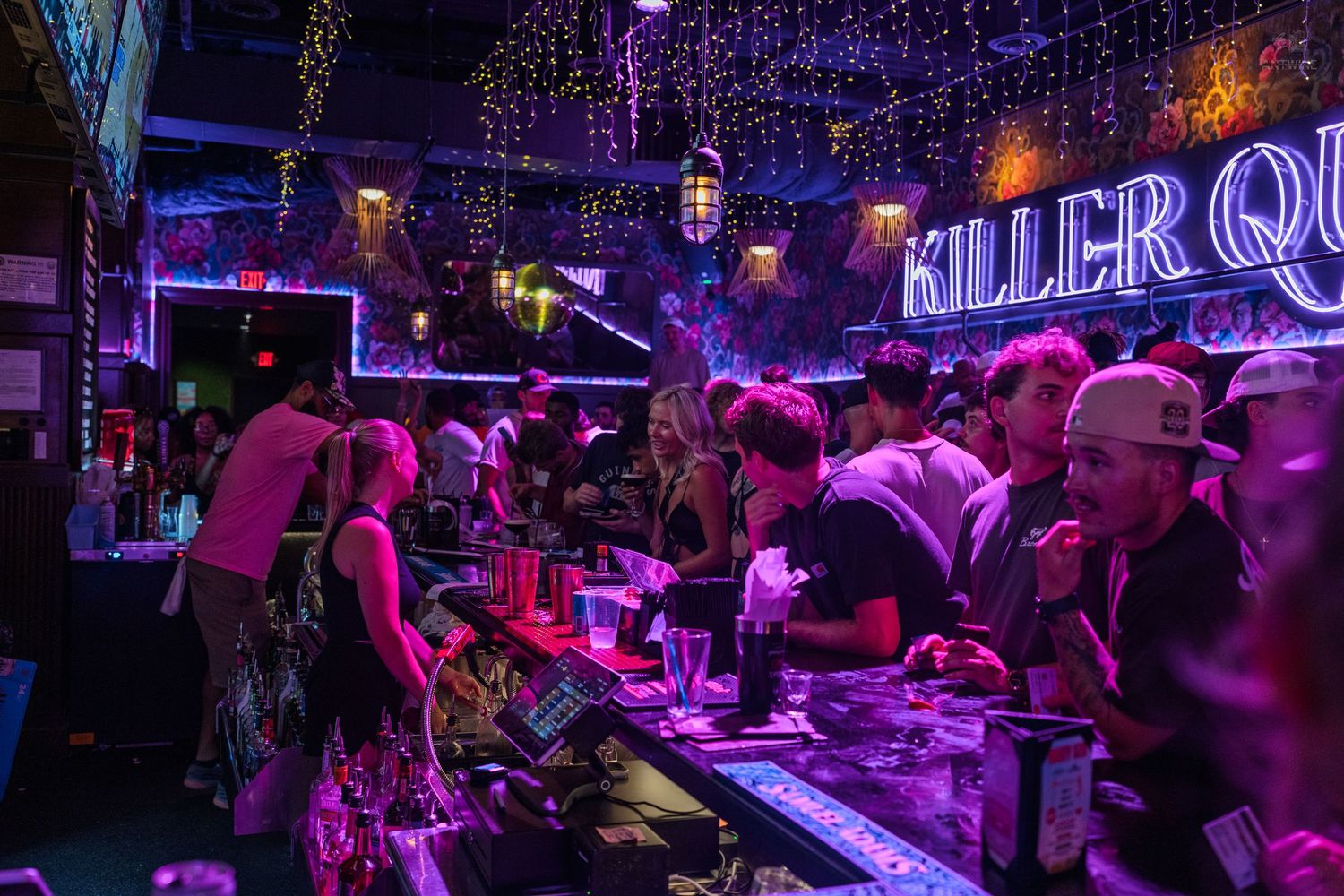 Crowded neon-lit bar with purple lights and Killer Queen sign, patrons socializing at counter.
