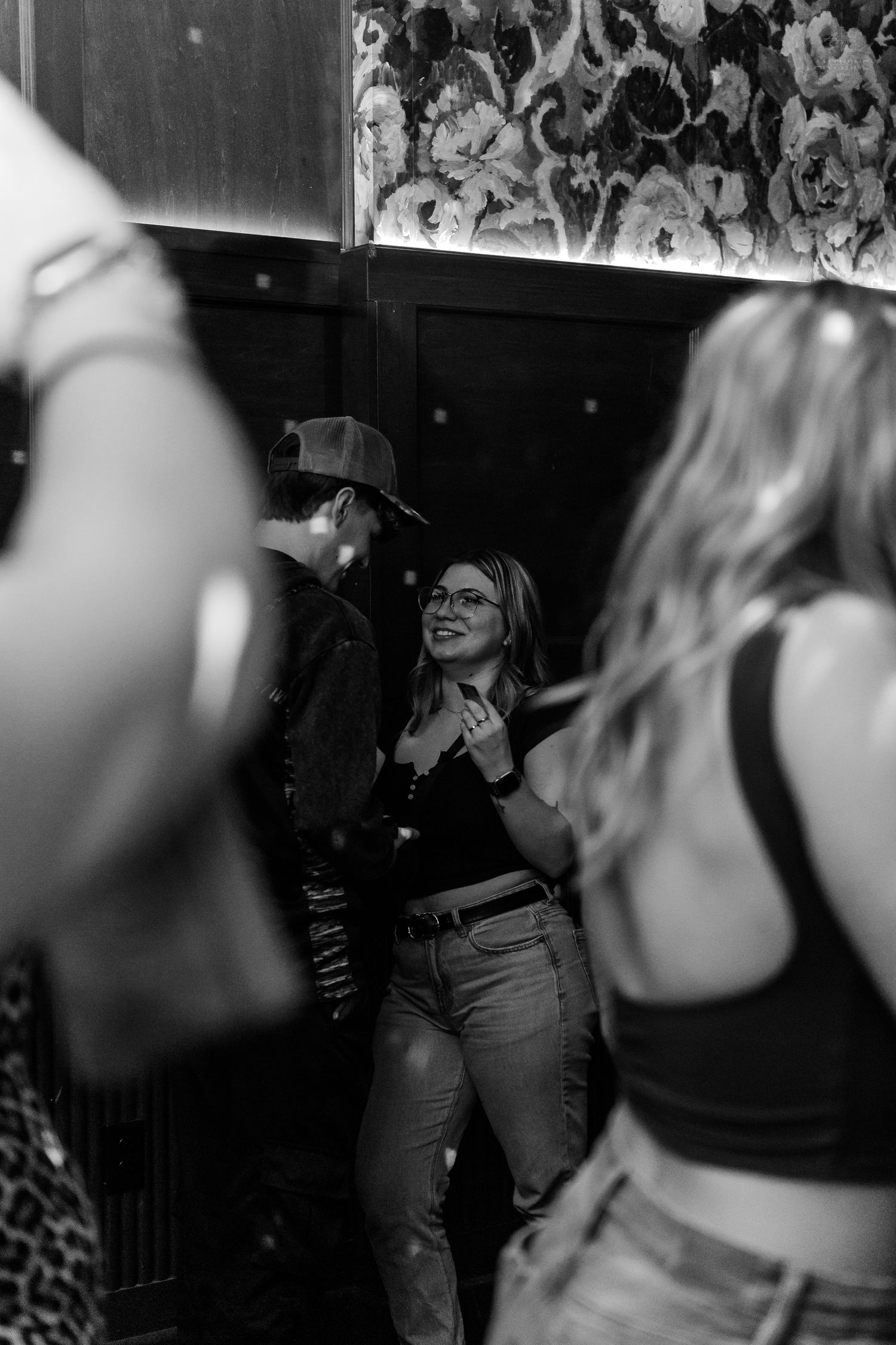 Black and white photo of a smiling woman in glasses dancing at a nightclub with floral wallpaper.