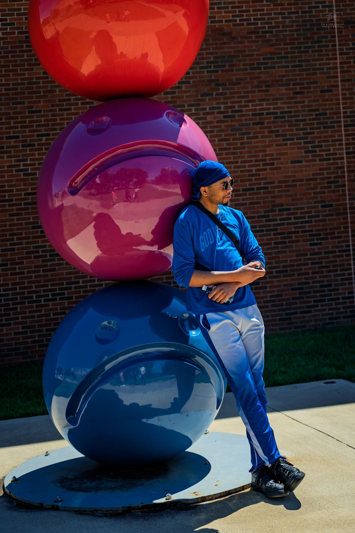 Man in blue outfit resting against bold red, purple, and blue stacked sphere sculpture outdoors.