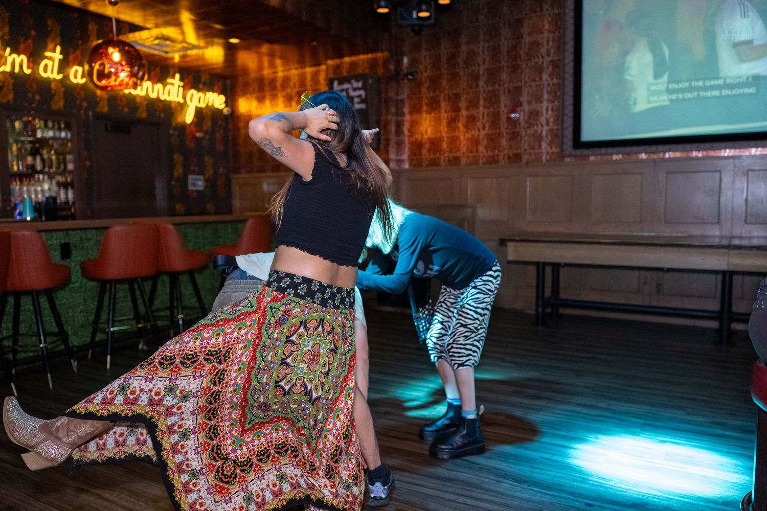 Woman in colorful maxi skirt poses while photographer crouches to take photo in a bar setting.