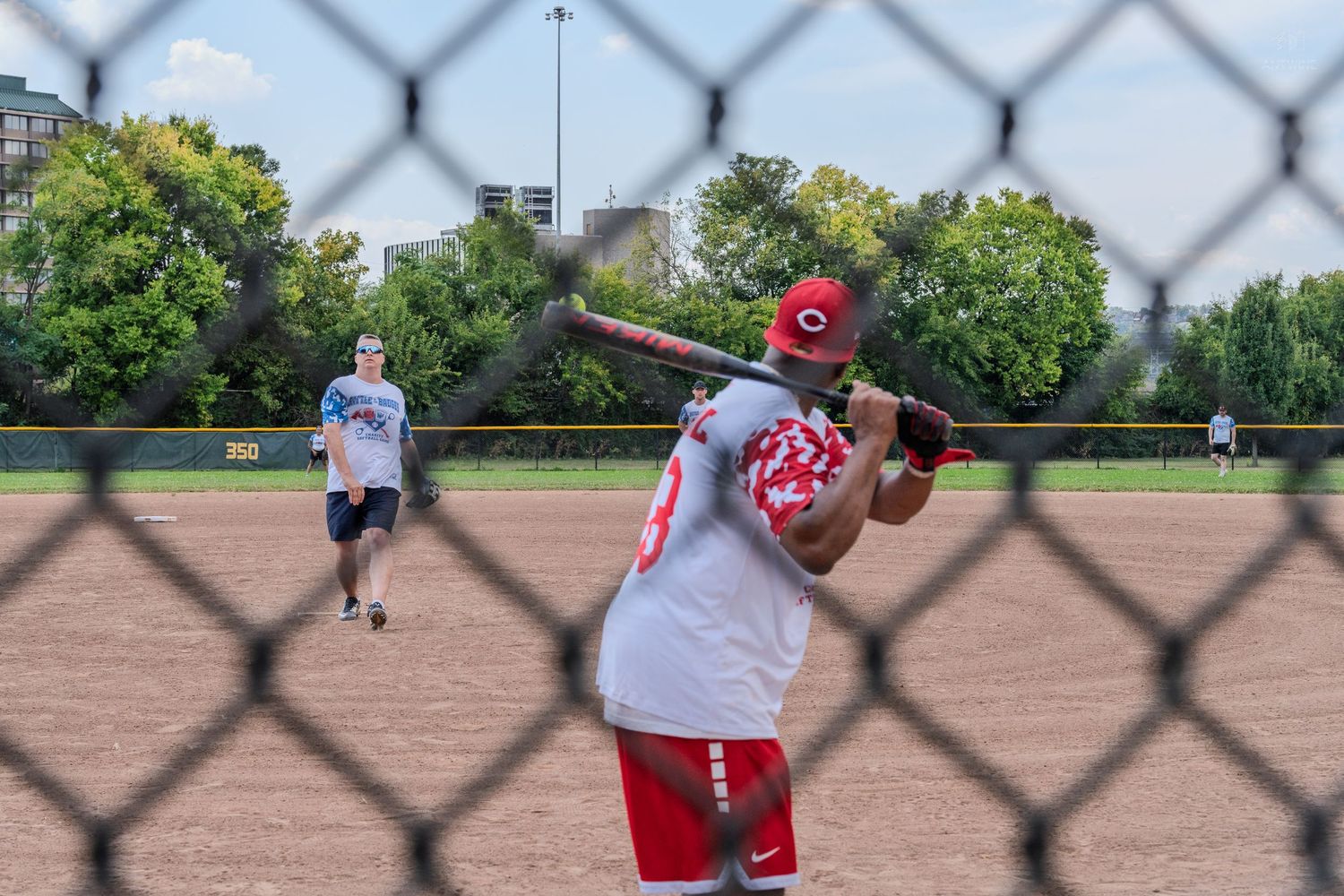 Softball batter in red uniform preparing to swing, viewed through a chain-link fence on a sunny day.