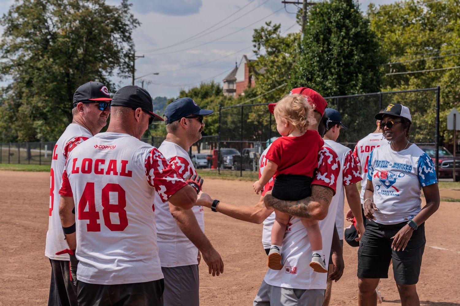 Softball players in Local 48 jerseys gather around a child in red on a sunny outdoor field.