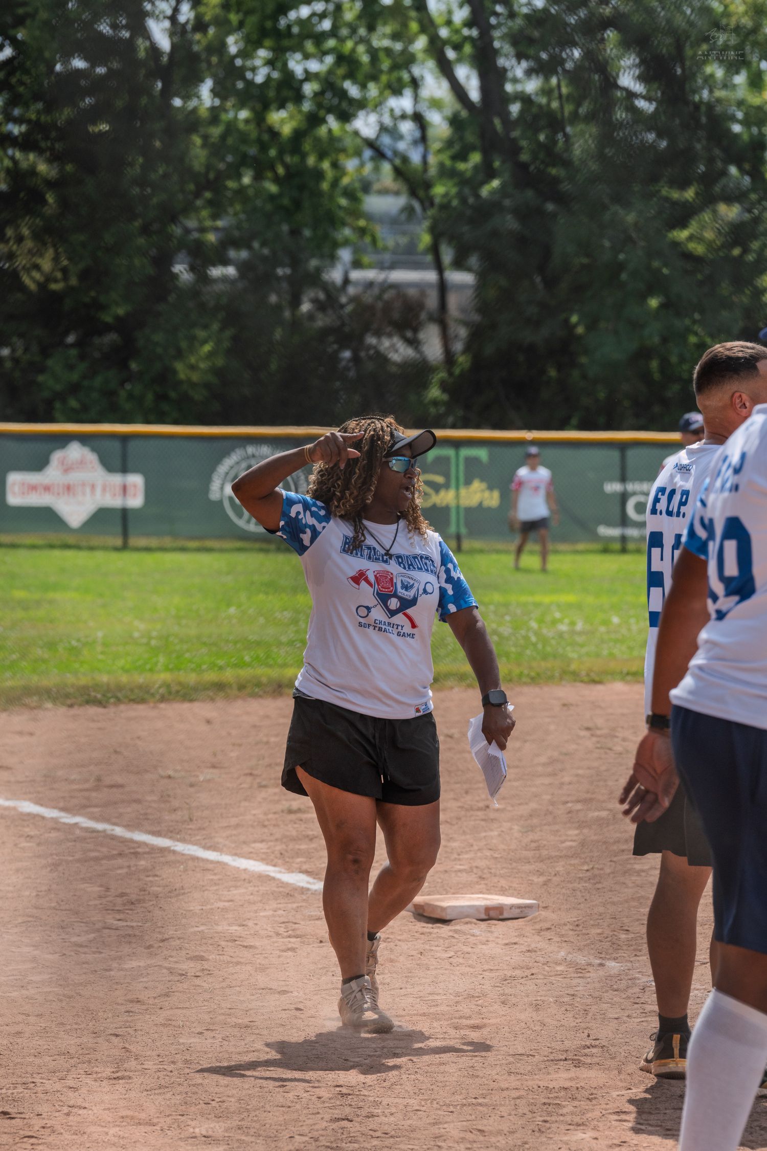 Woman in softball uniform adjusts hat on base during outdoor charity softball game on sunny day.