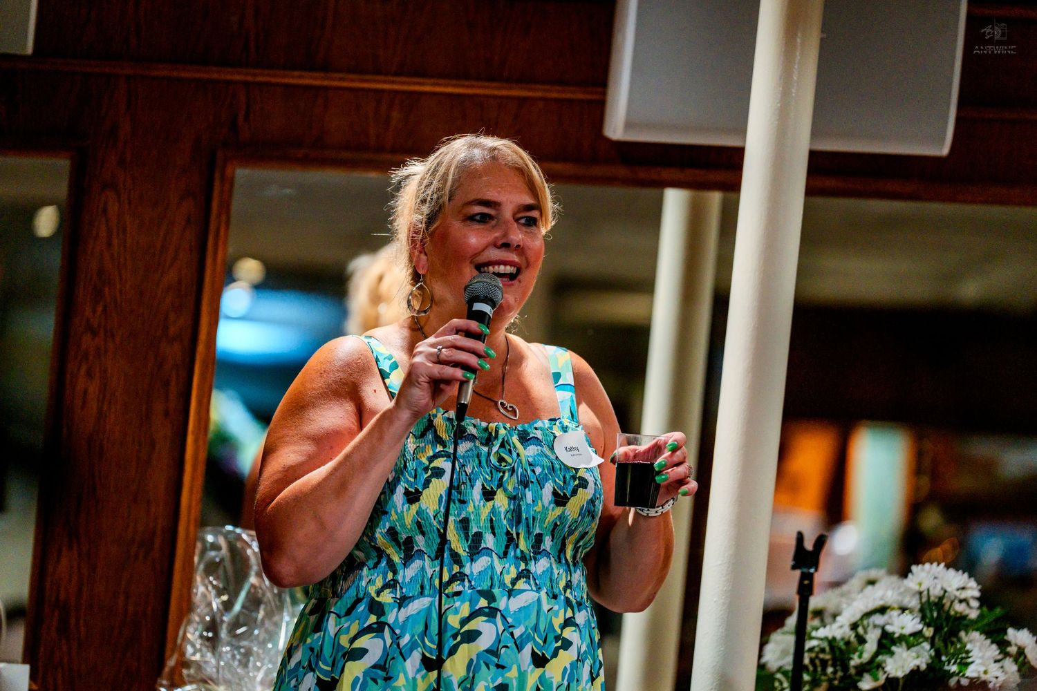 Woman smiling and speaking into microphone at indoor event wearing colorful floral dress.