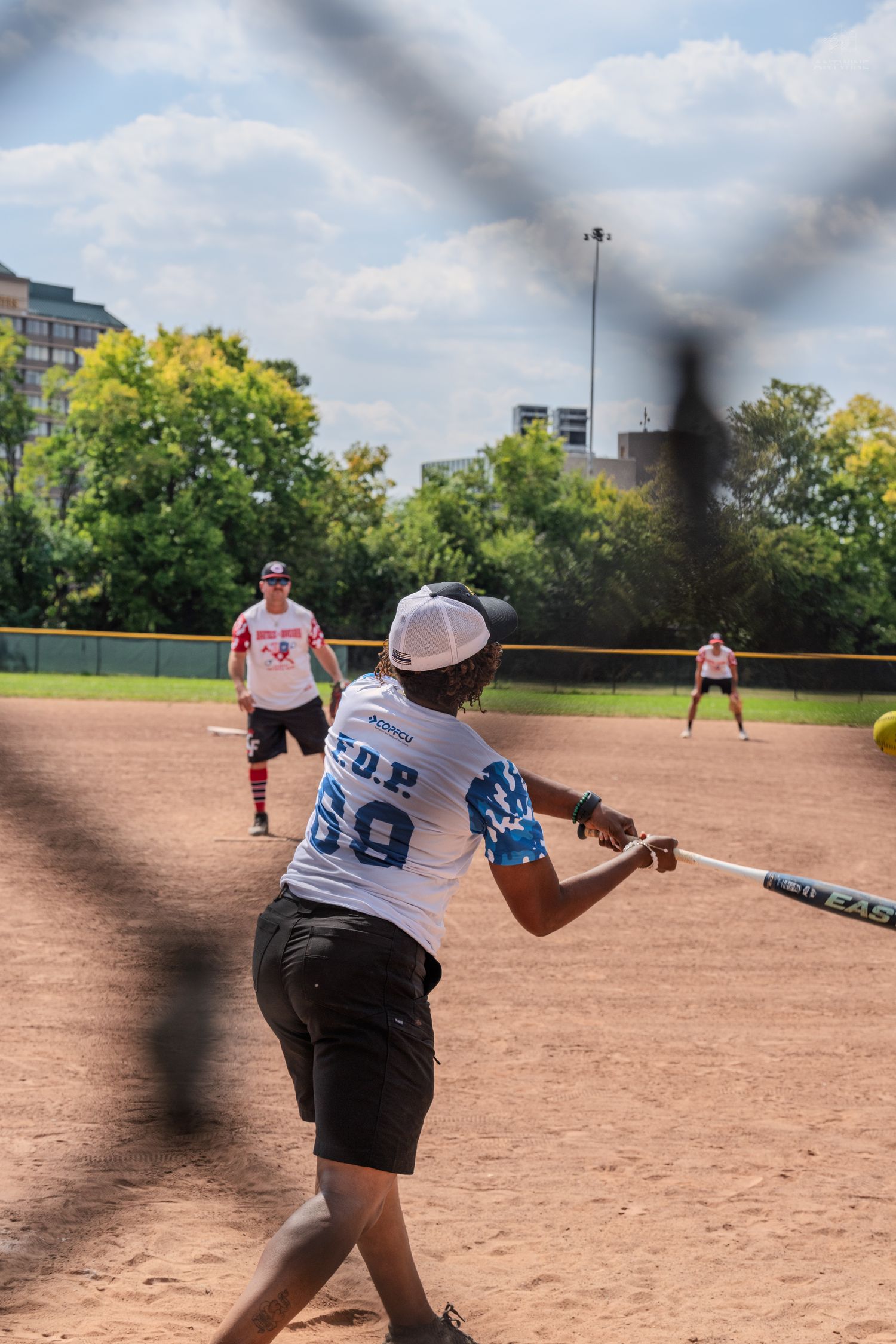 Softball batter in F.O.P. jersey #69 swings at a pitch during an outdoor game on a dirt field.
