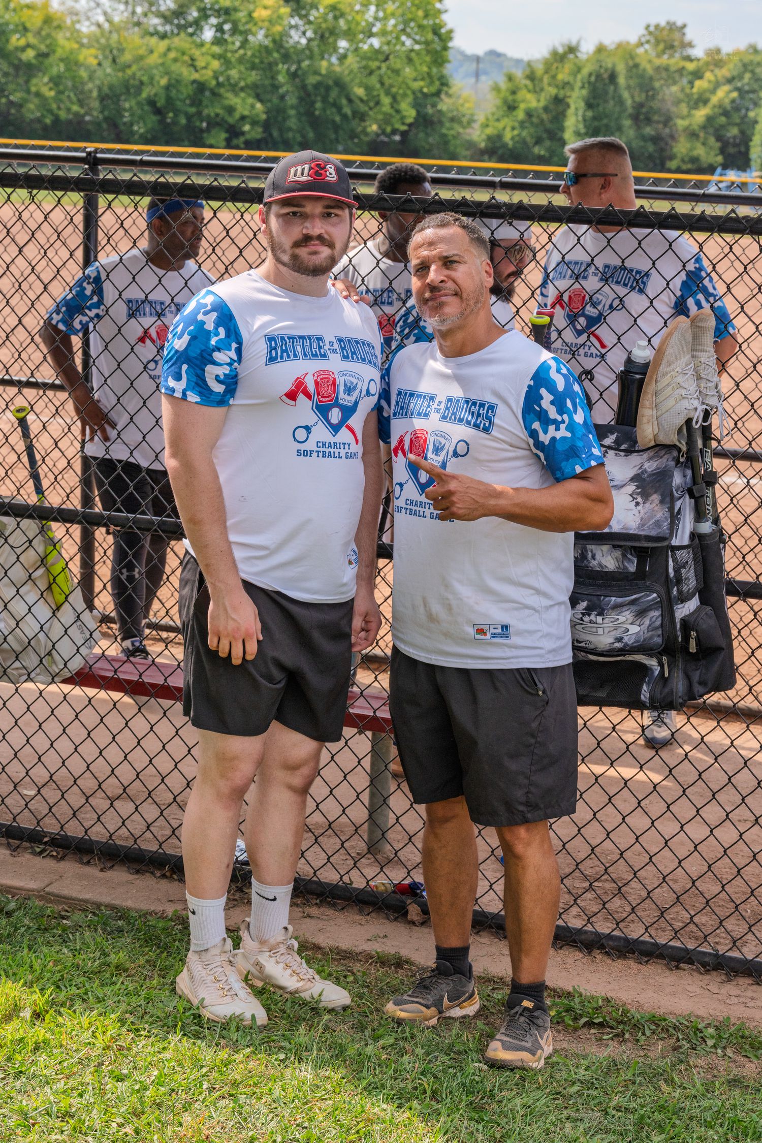 Two men in matching Battle of Badges charity softball game jerseys pose together near chain-link fence.