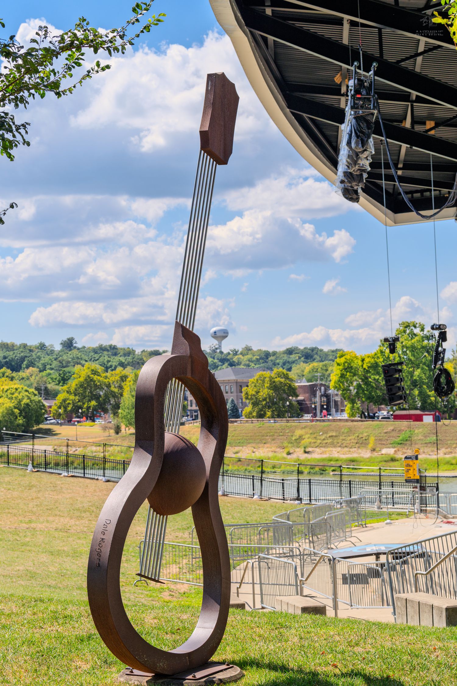 Large metal guitar sculpture stands outdoors near an amphitheater stage with green trees and blue sky backdrop.