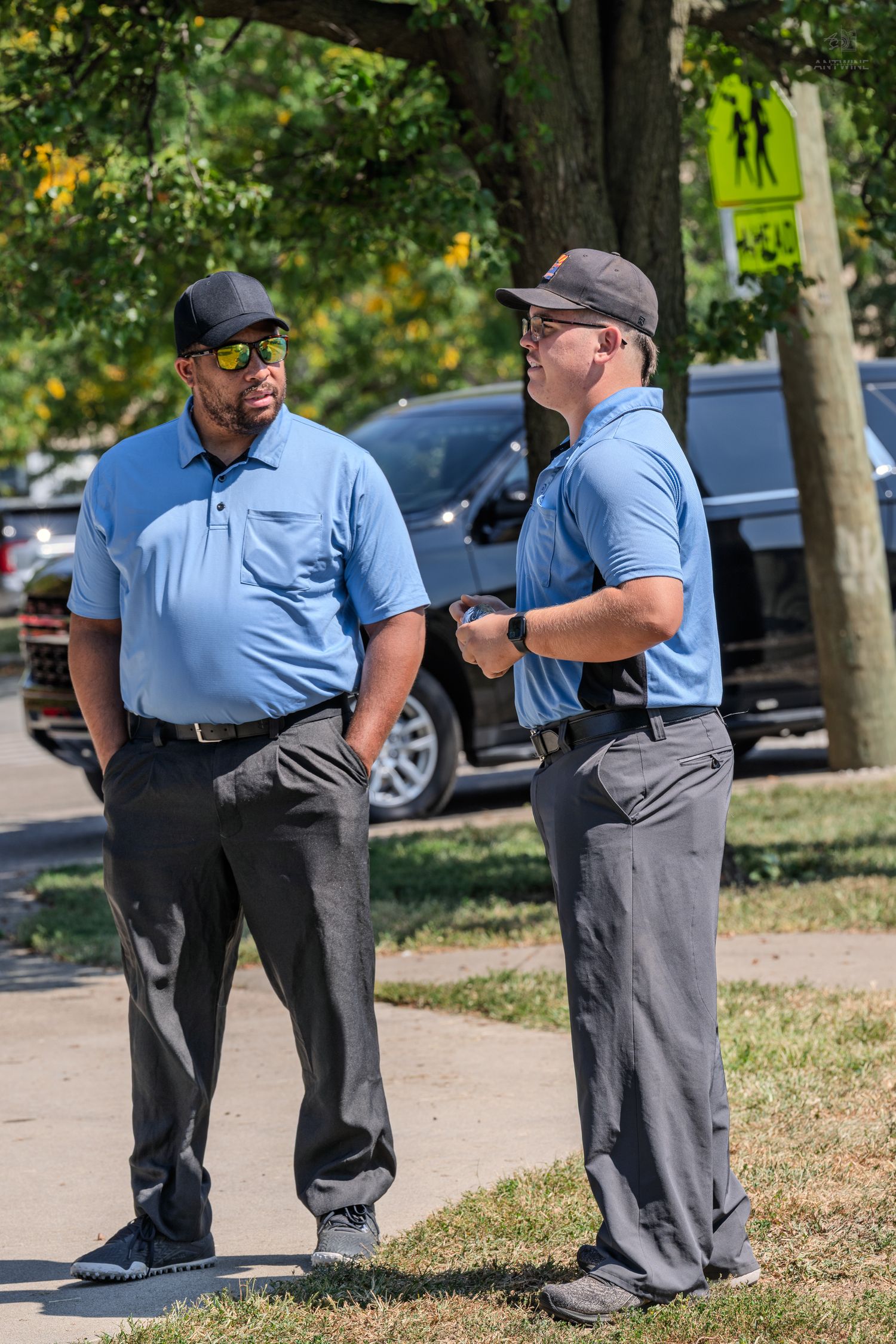 Two men in matching blue polo shirts and caps having a conversation on a tree-lined sidewalk.
