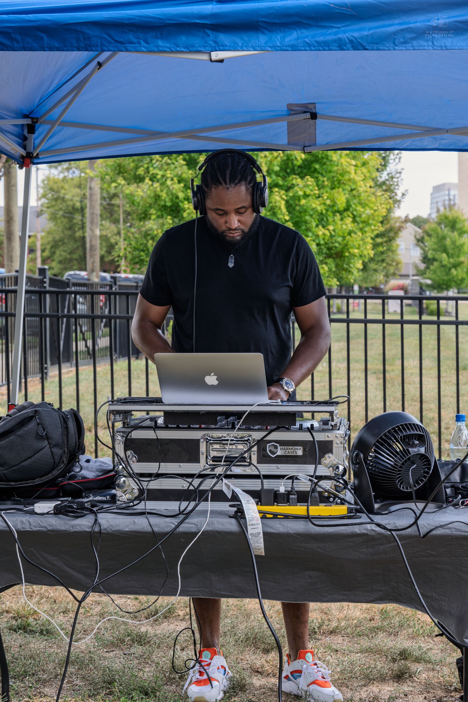A DJ wearing headphones operates a MacBook laptop and mixer under a blue canopy tent outdoors.