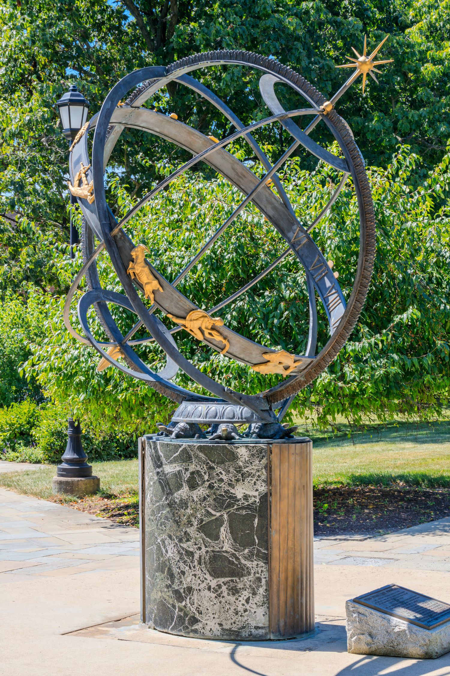 Ornate armillary sphere sundial on marble pedestal with golden figures, set in a sunny park with lush green trees.