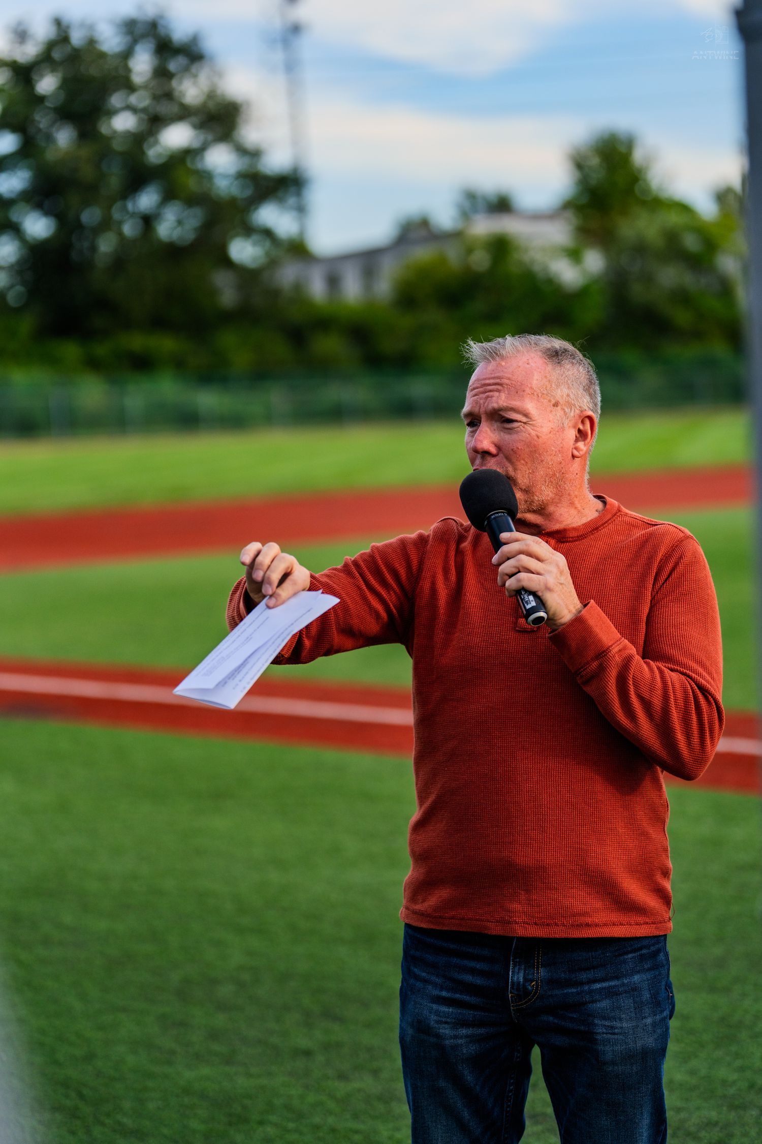 Middle-aged man in red sweater speaking into microphone at outdoor sports track, holding papers.