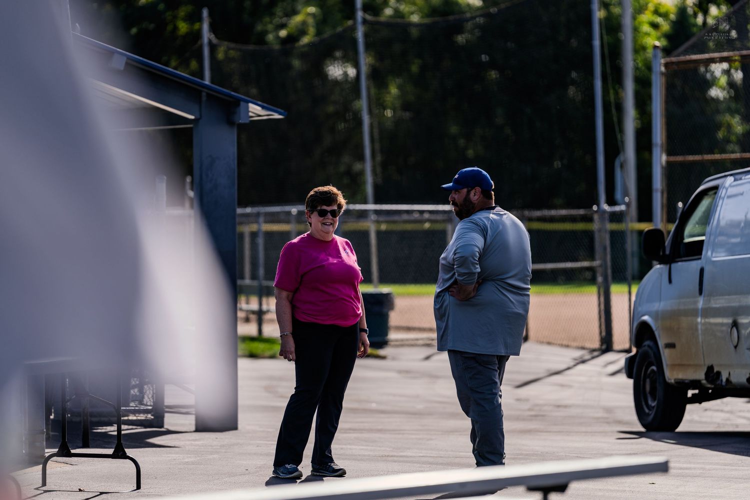 Two people conversing outdoors near a parking lot on a sunny day, woman in pink shirt and man in blue shirt.