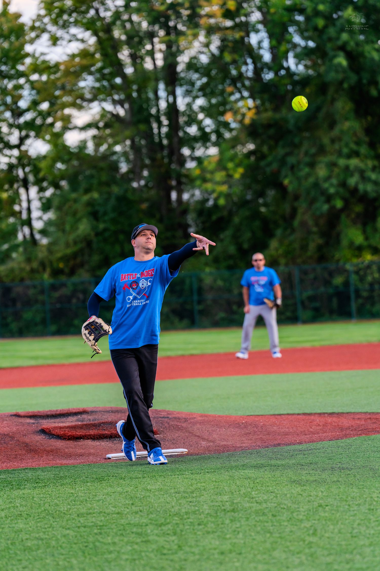 A softball pitcher in a blue shirt releases a yellow ball on a well-maintained baseball field at dusk.