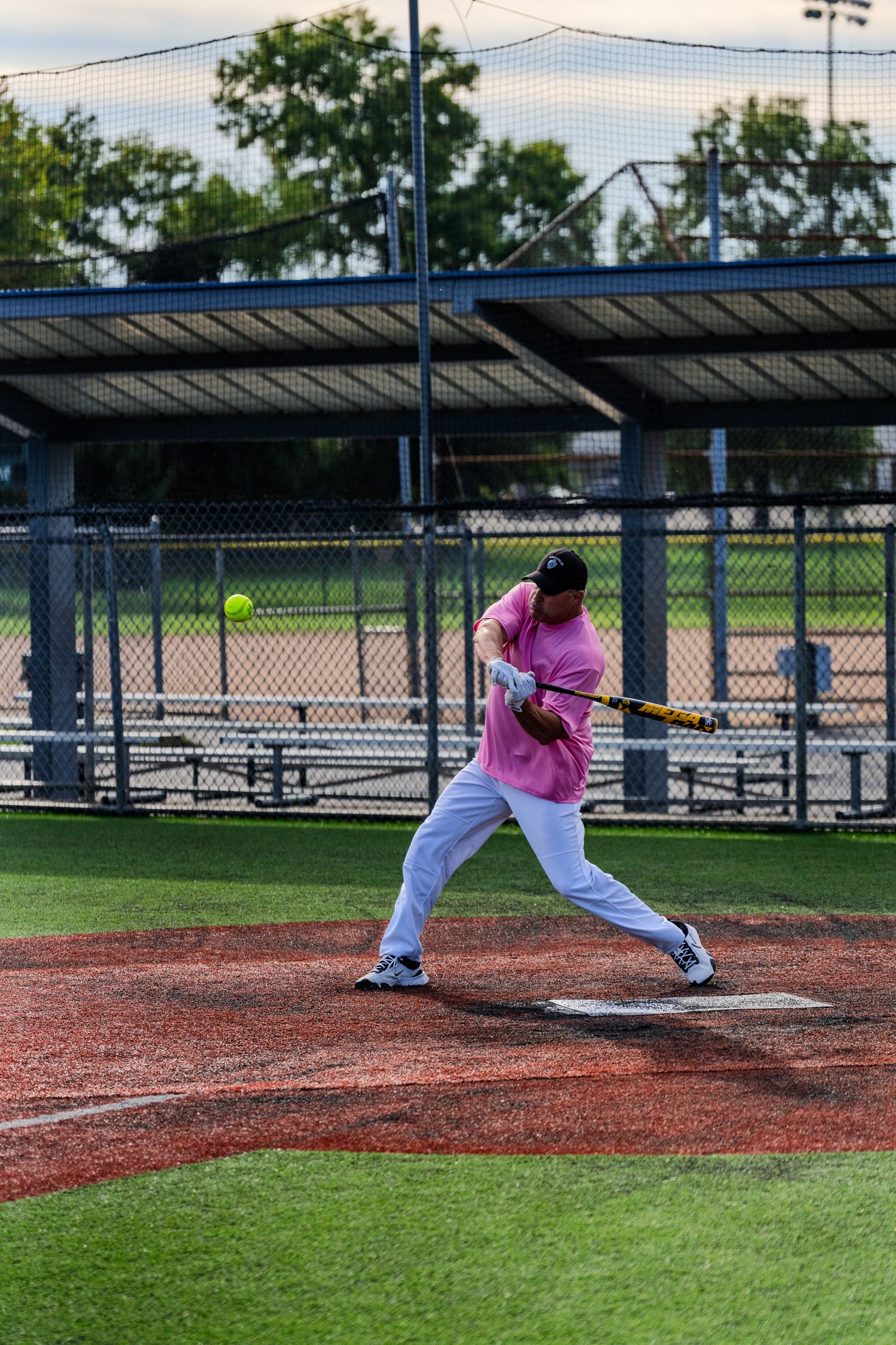 A batter in a pink shirt swings at a yellow softball during a recreational game on a red dirt field.