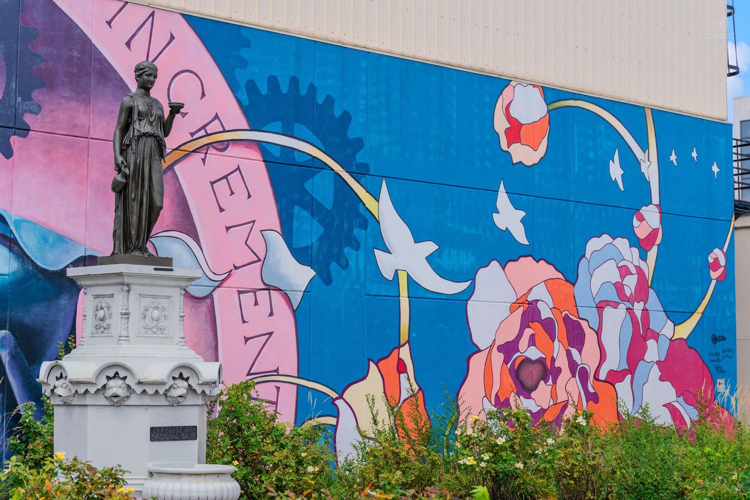 Colorful street art mural with pink, blue and orange flowers behind a classical stone statue on a city building.