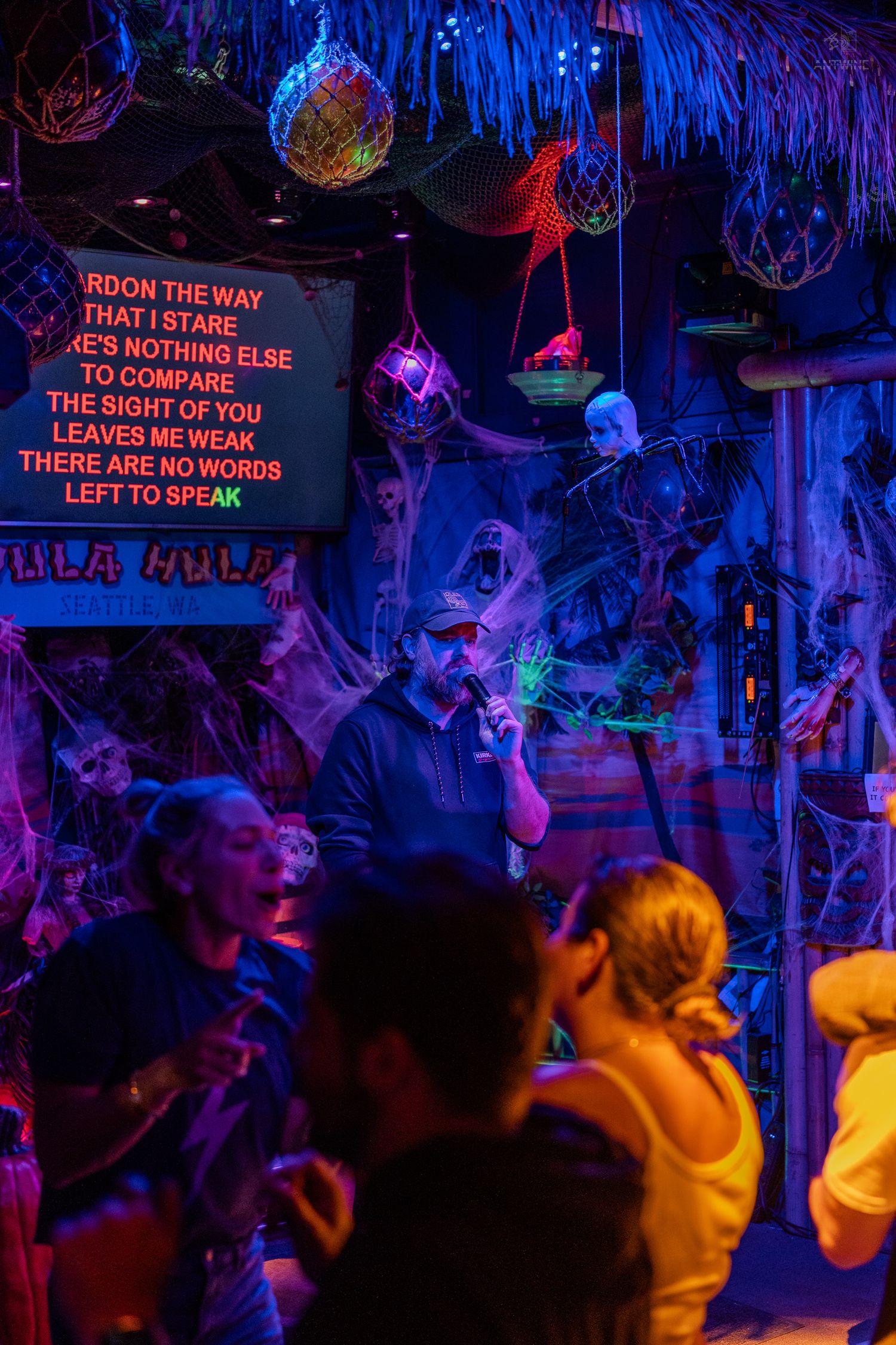 Man singing karaoke at Halloween-themed Hula Hula bar surrounded by spooky neon decorations.