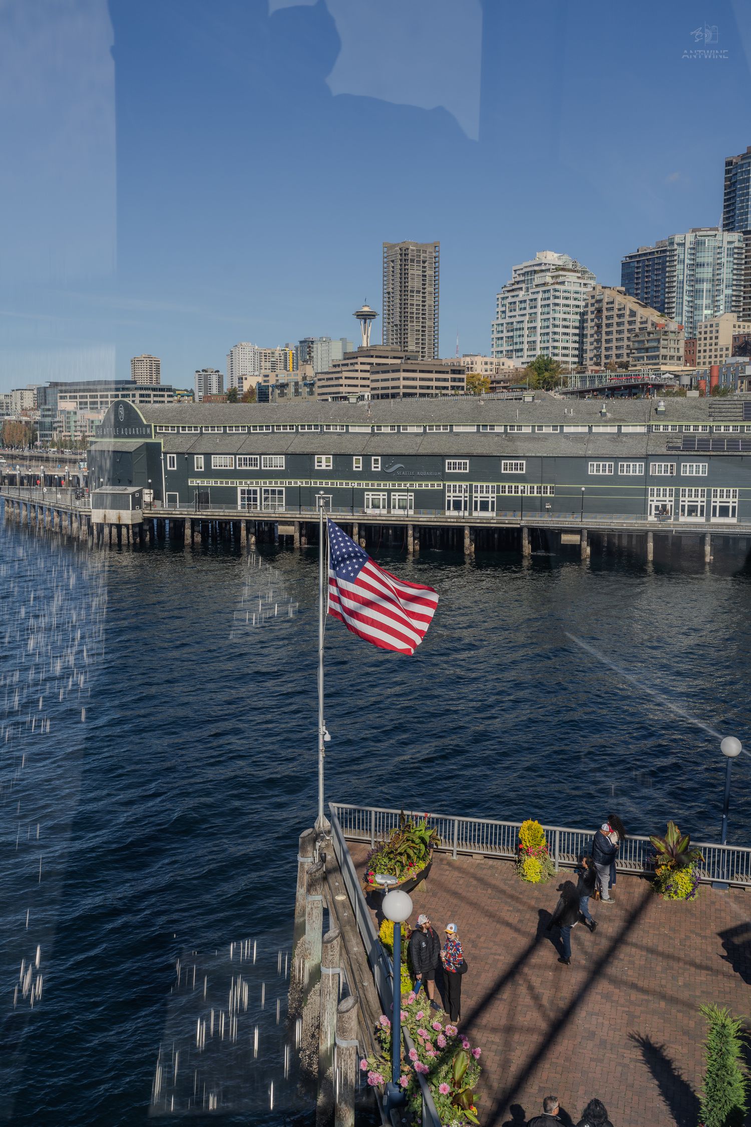American flag flies over Seattle waterfront pier with the Space Needle and city skyline visible across Elliott Bay.