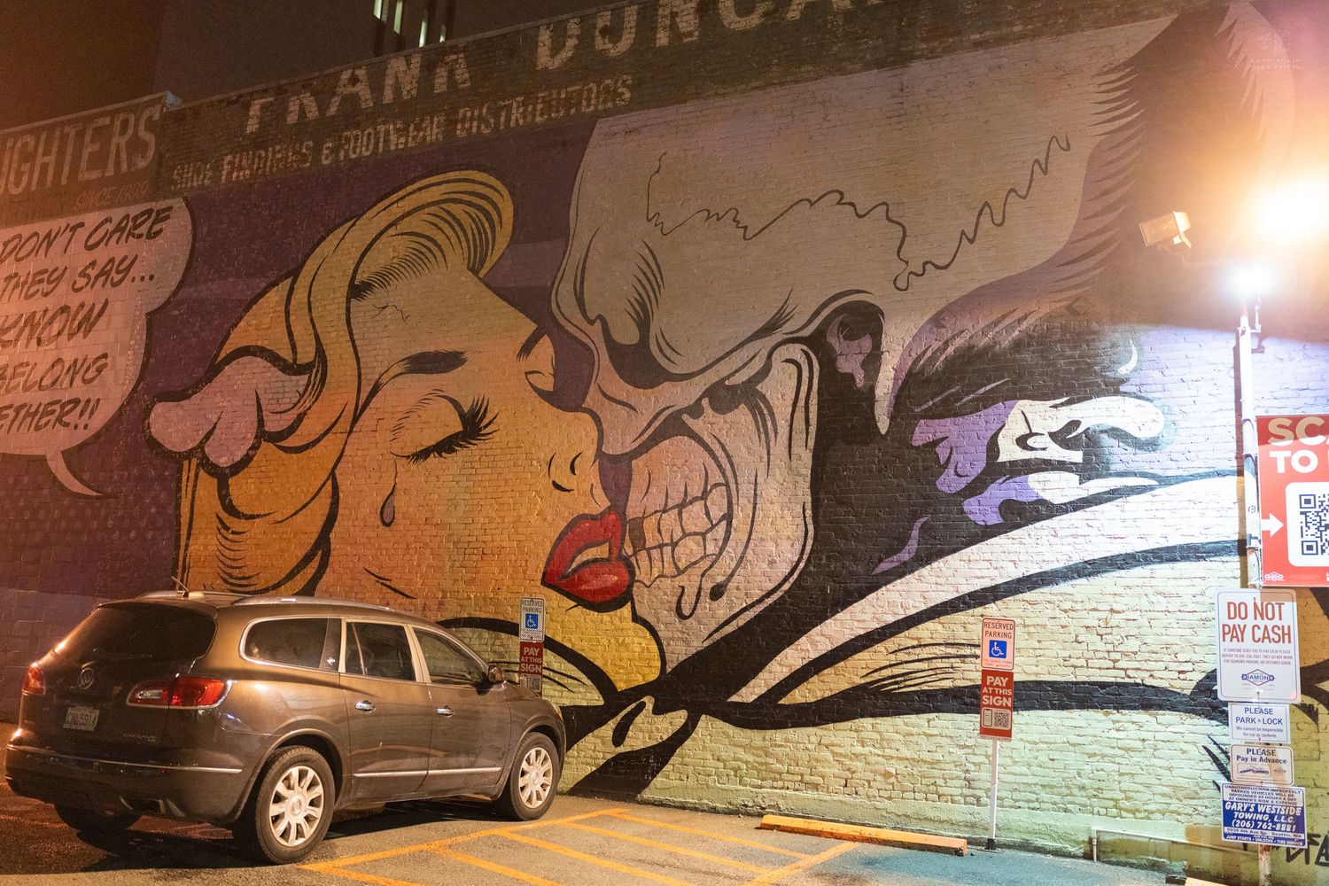 Colorful street art mural of a woman and skull on a brick wall at night with a parked car in foreground.