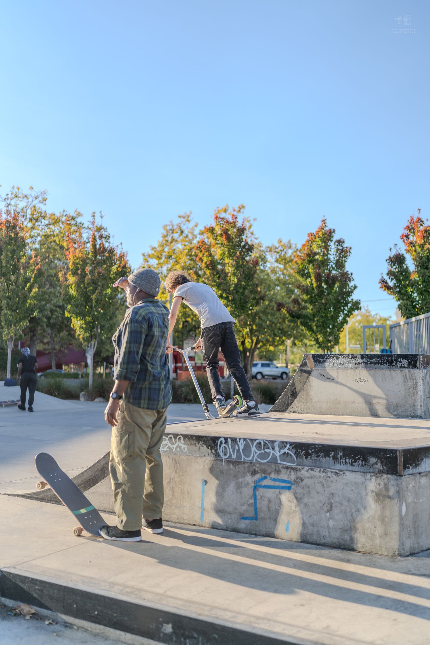 Skateboarders practice tricks at an outdoor skate park with colorful autumn trees in the background.